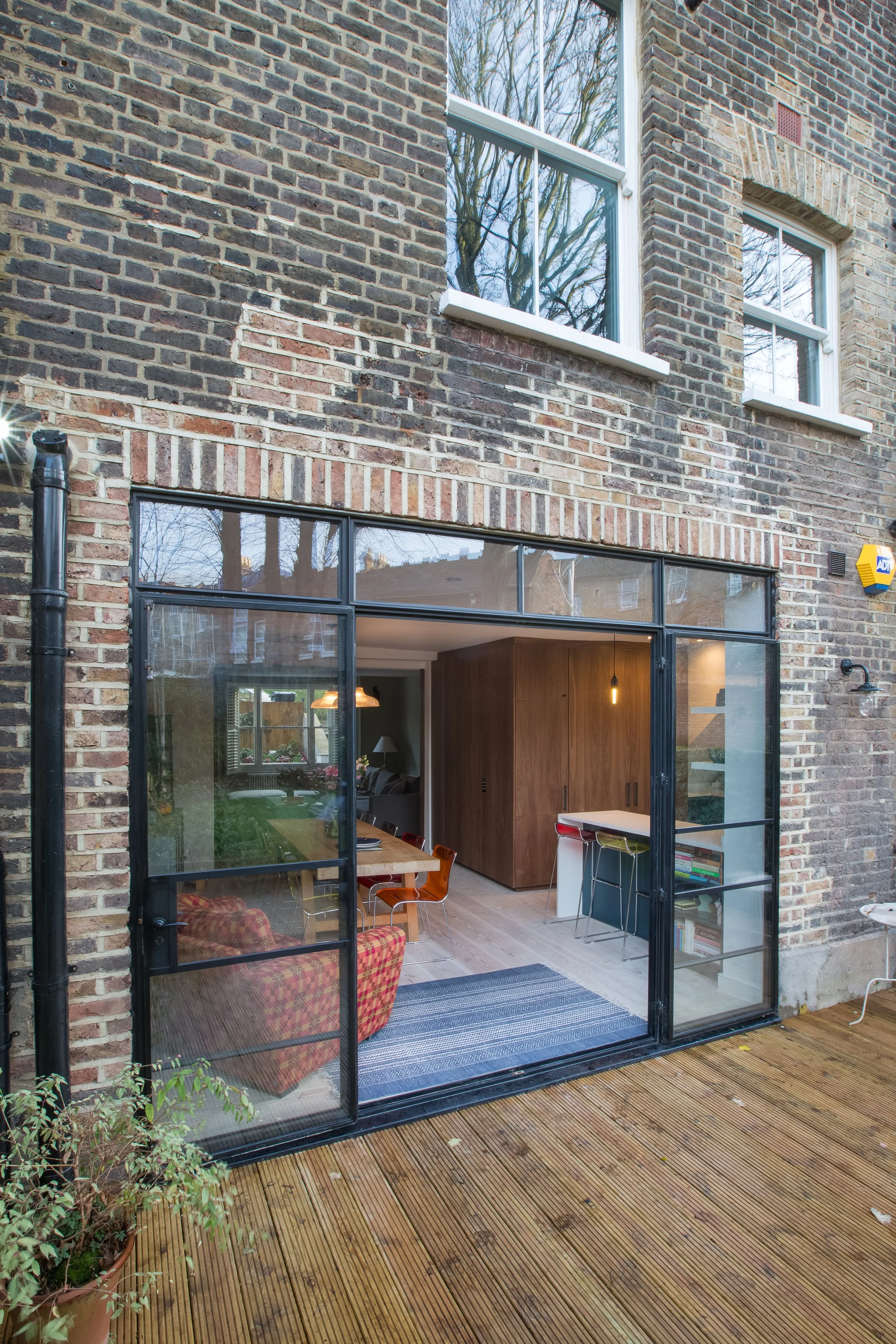A brick house with large sliding glass doors opening to a wooden deck. Inside, a modern kitchen and living space are visible through the glass, with natural light streaming in. The house has two upper-story windows with white frames and a small vent.