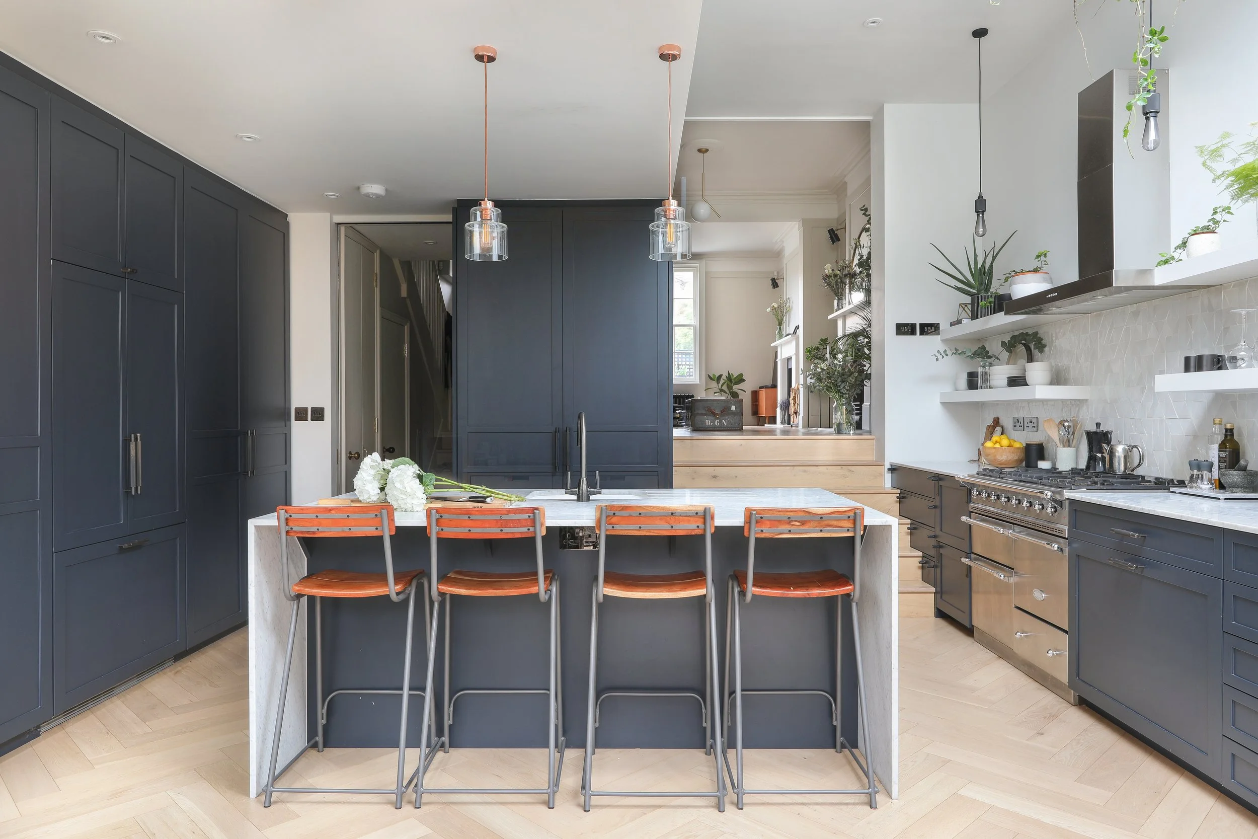 Modern kitchen with navy blue cabinets, white countertops, and a central island with orange bar stools. Open shelving with plants and kitchenware, stainless steel oven, and pendant lights hanging above the island.