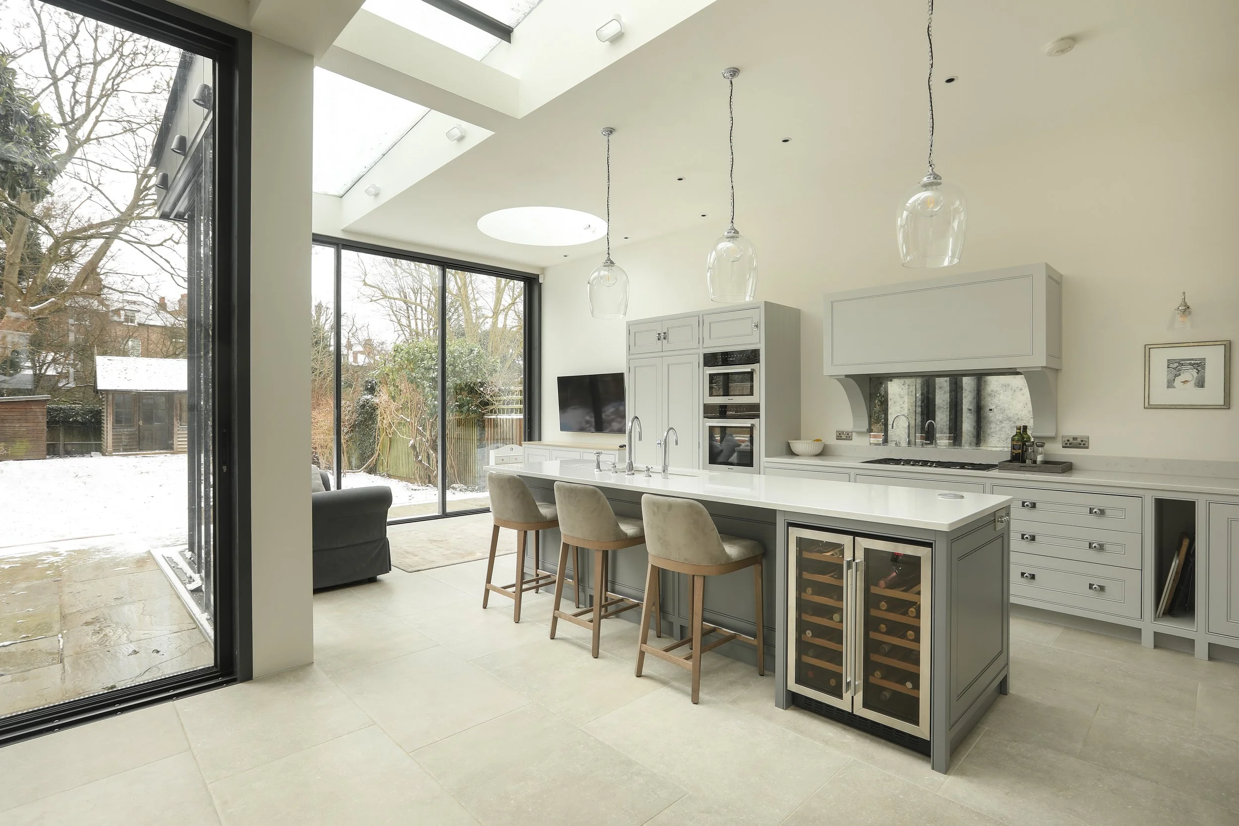 Modern kitchen with large windows and sliding glass door, white countertops, gray cabinetry, hanging glass pendant lights, and a view of snowy backyard outside.
