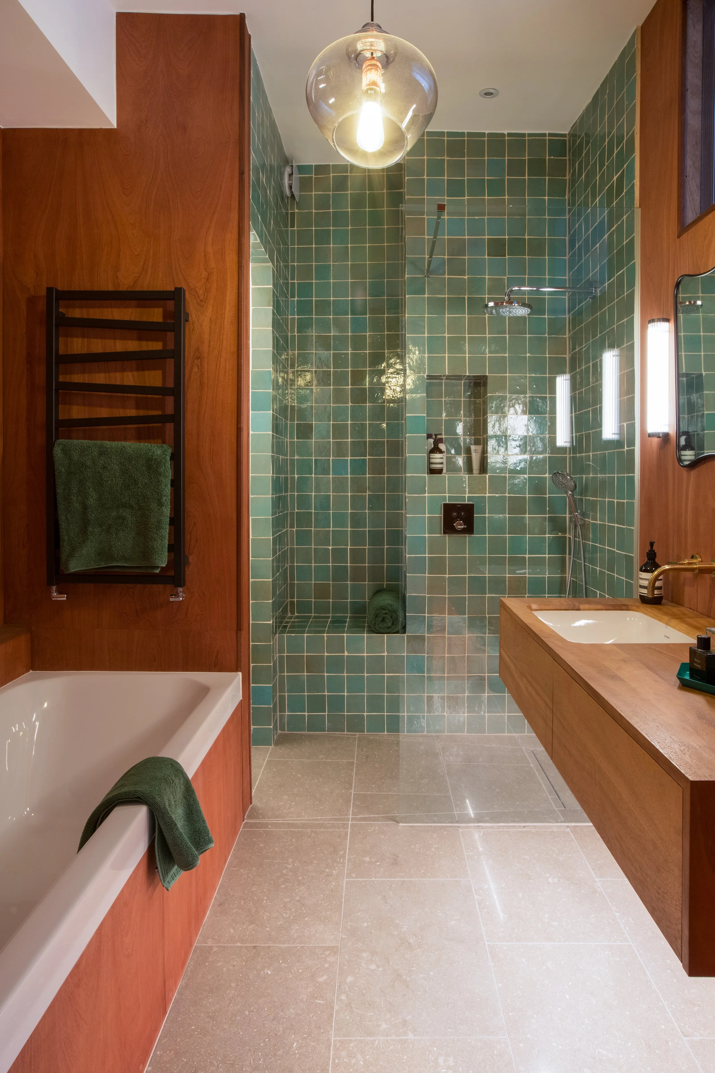 Modern bathroom with a green tiled walk-in shower, wooden vanity with a white sink, and a bathtub with a green towel. Wooden accents and a hanging glass light fixture complete the decor.