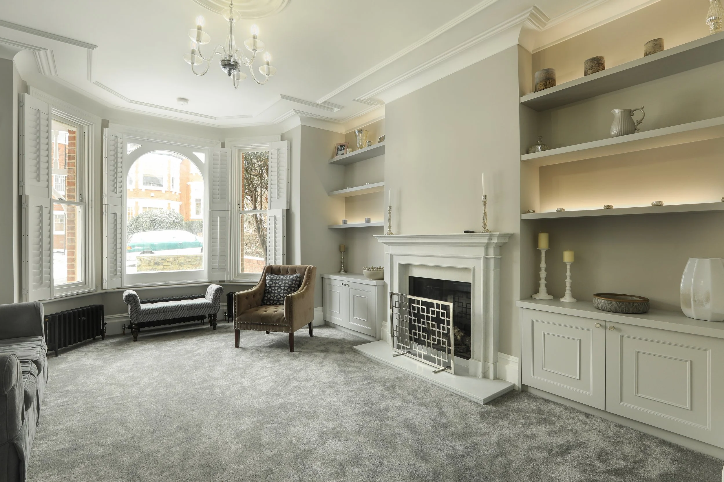 Living room with large bay windows, white shutters, beige walls, and built-in shelves with decorative items. There's a white fireplace with candles and candlesticks. Furnishings include patterned armchairs and a gray carpet.