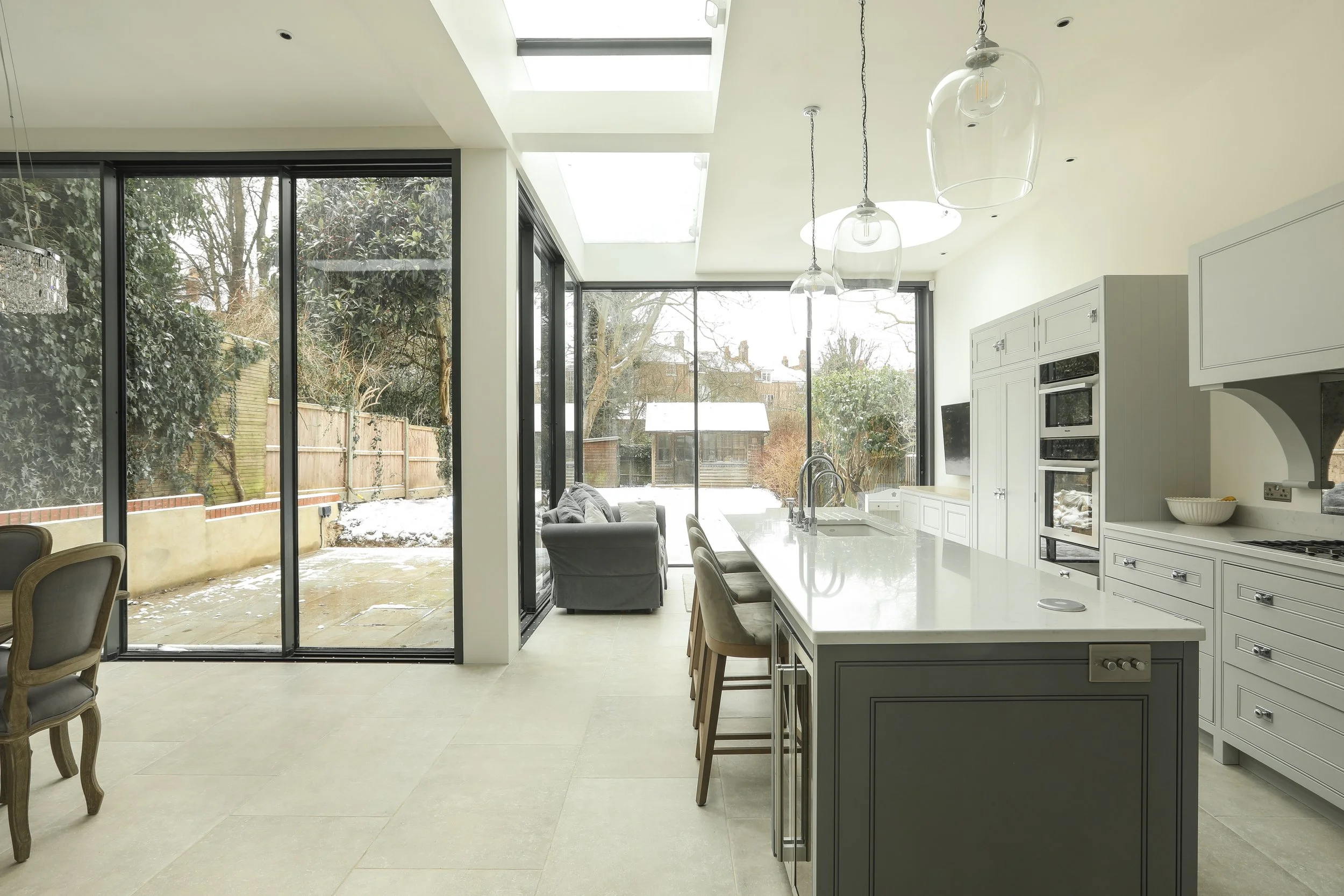 Bright, modern kitchen with large windows and sliding glass doors showing a snowy backyard. White cabinets, a kitchen island with beige chairs, and pendant lights hanging from the ceiling.