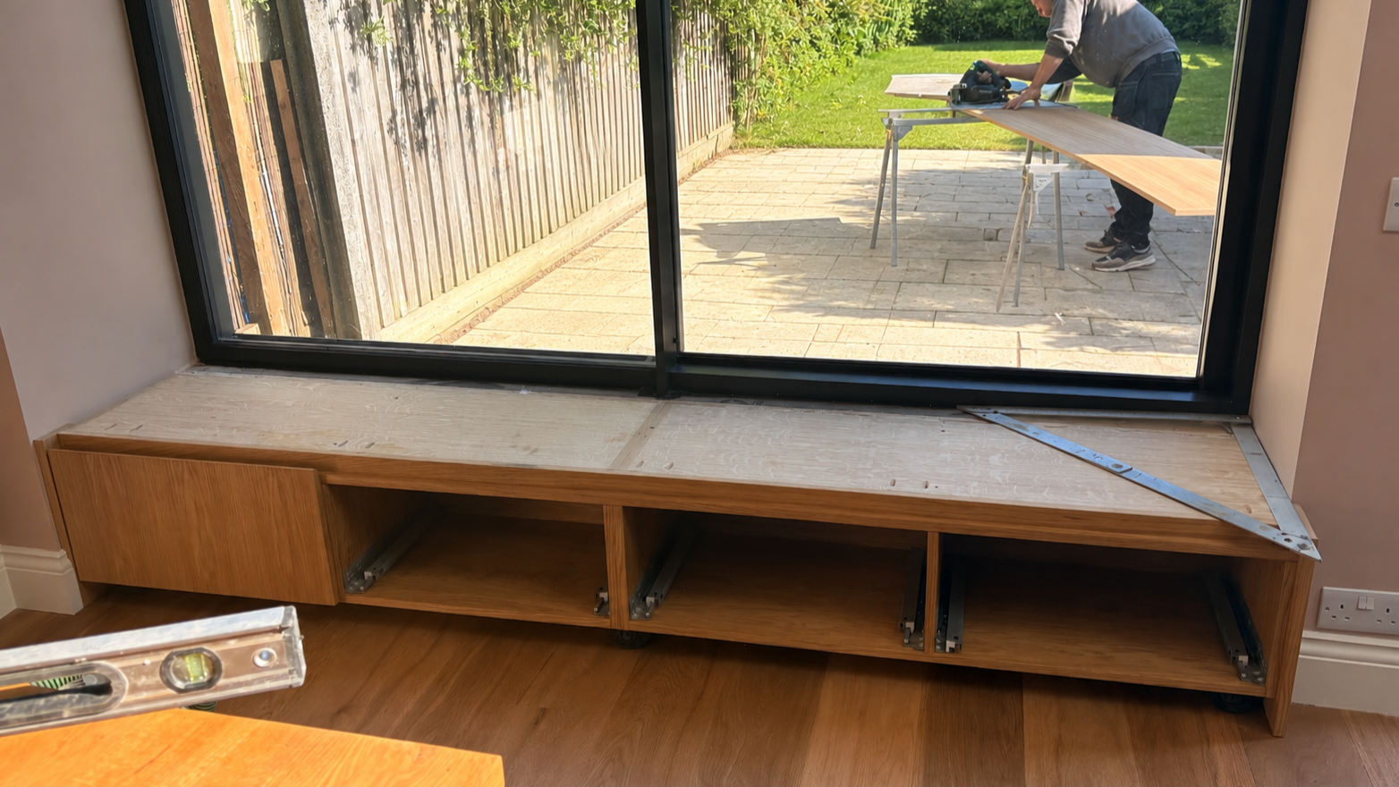 Wooden storage unit under construction in front of a large glass sliding door, with a person working outside cutting a wooden board.