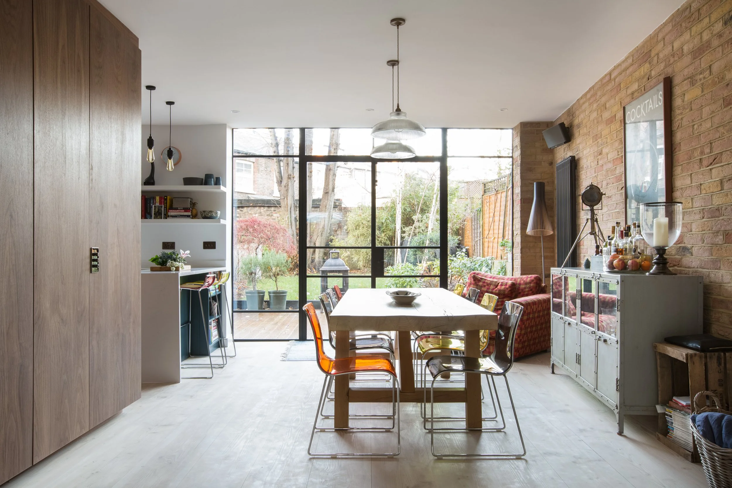 Interior of a modern dining room with a wooden table, colorful chairs, a brick accent wall, large windows, and various decorative items.