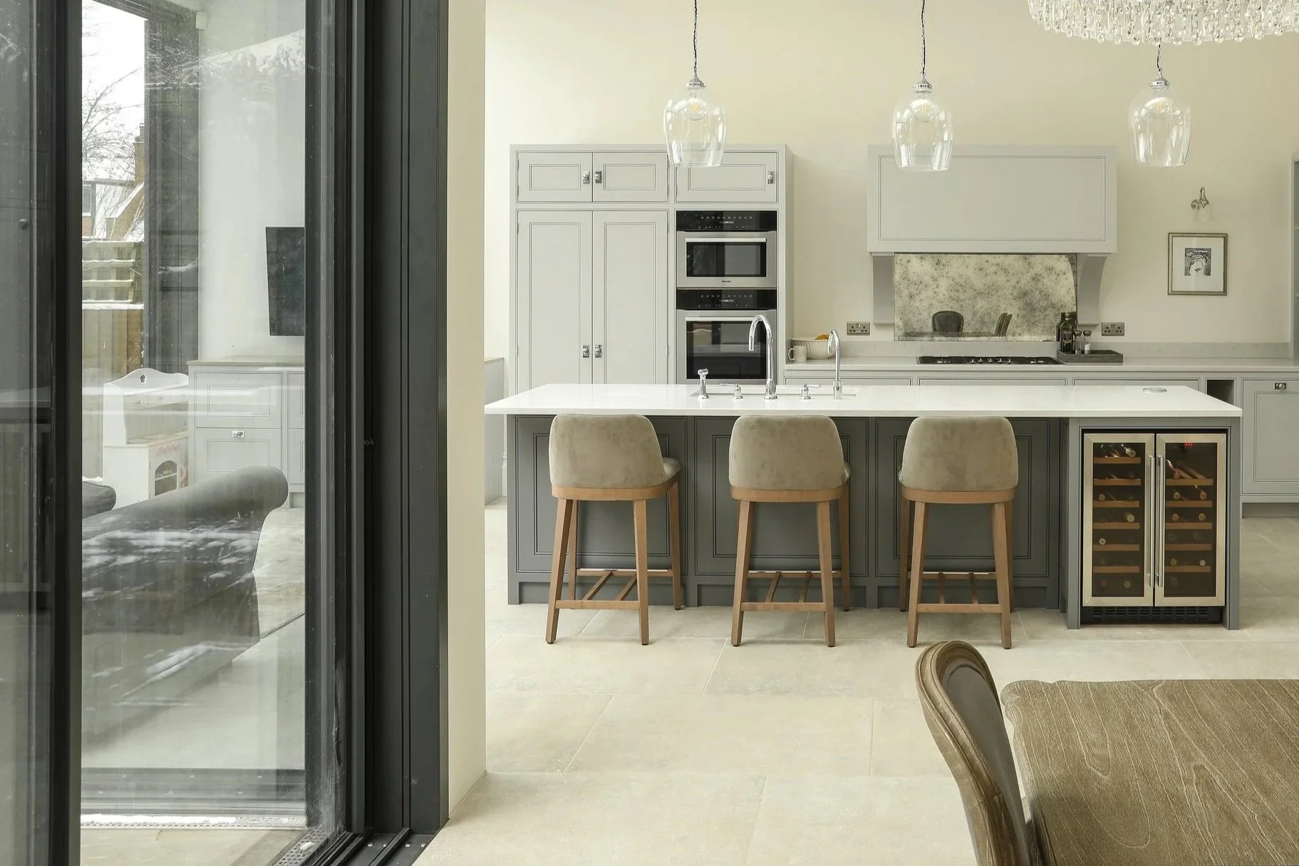 Modern kitchen with white cabinets, a white kitchen island with beige barstools, stainless steel appliances, a large crystal chandelier, pendant lights, a wall clock, and sliding glass doors leading outside.