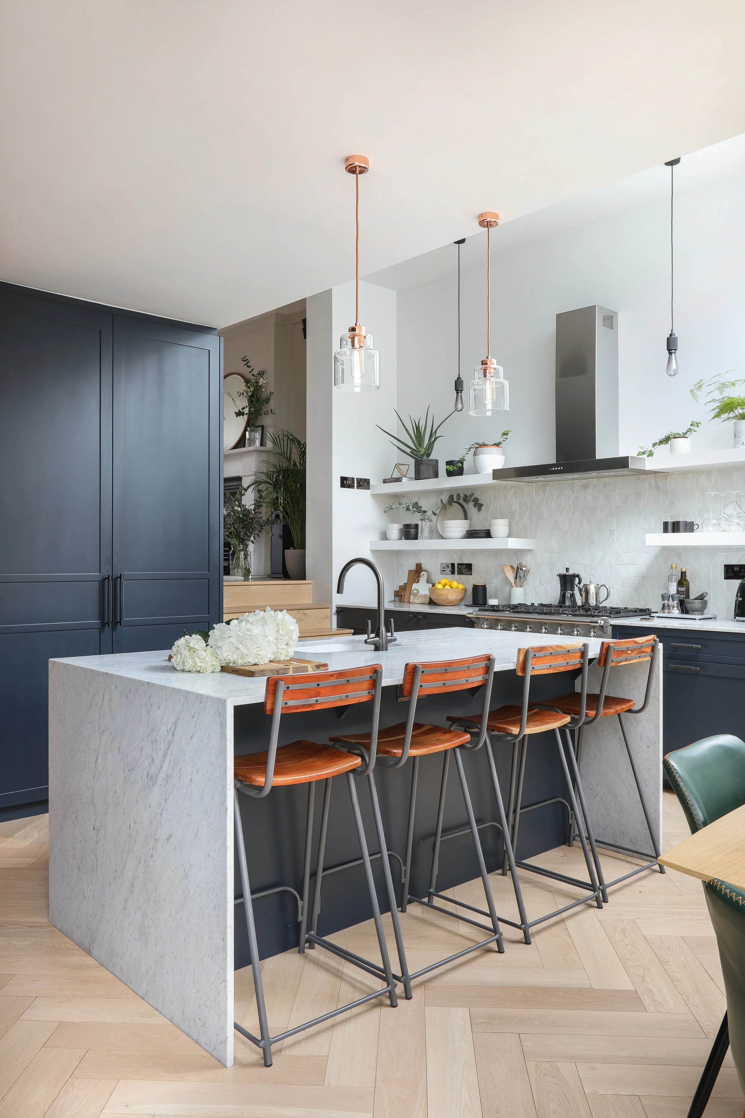 Modern kitchen with a large white island, navy blue cabinets, open shelving with plants and dishes, pendant lights, and four bar stools with wooden seats.