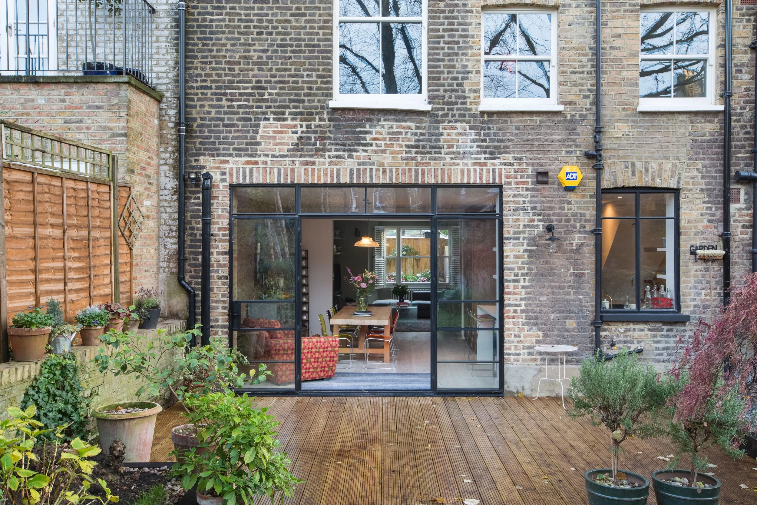 Backyard patio view of a house with large windows and a sliding glass door, with potted plants and a wooden deck in the foreground.