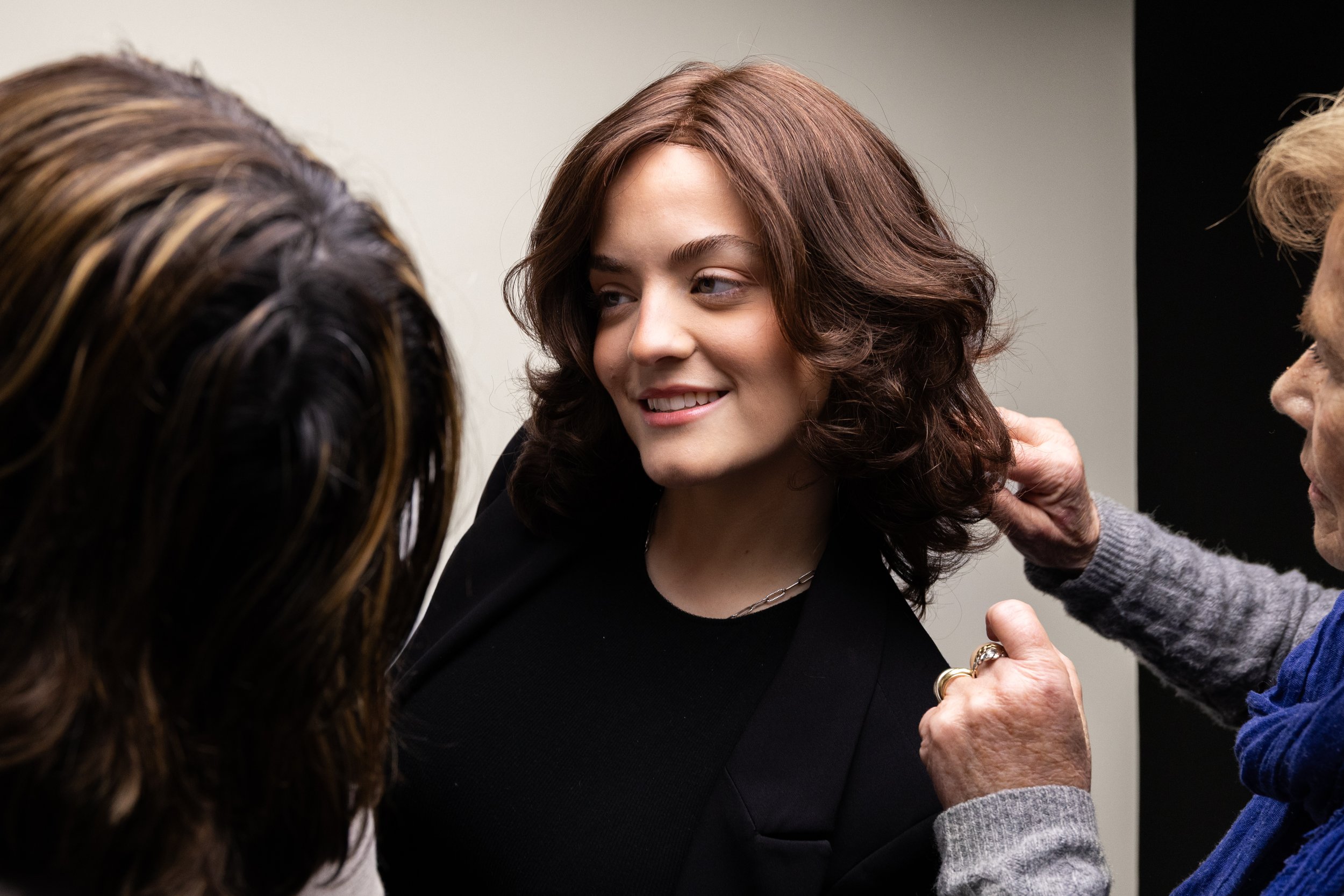 A woman with wavy brown hair smiling while being attended to by two people, one with long dark hair and the other with short blonde hair. The setting appears to be a backstage or salon environment.
