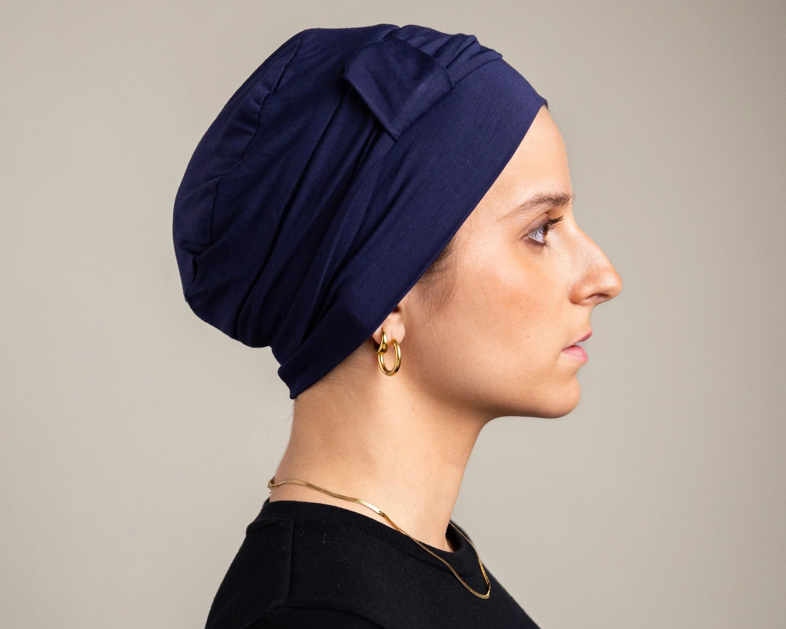 Profile of a woman with tan skin wearing a navy headscarf and gold jewelry, facing right against a plain background.
