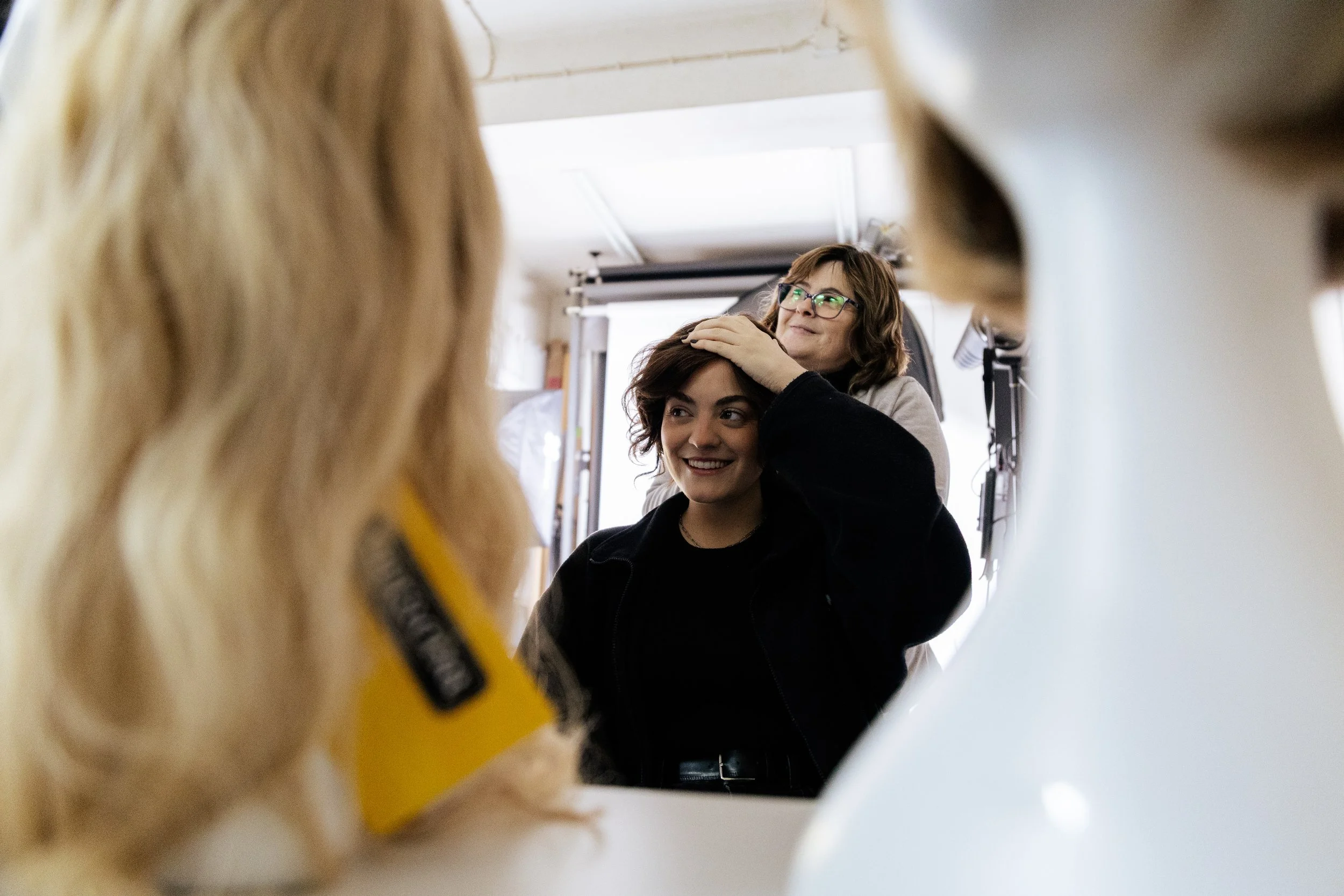 Two women look into a mirror, one with glasses and one with short dark hair, in a well-lit room. The photo is taken through a blurred foreground, possibly a person with long blonde hair.