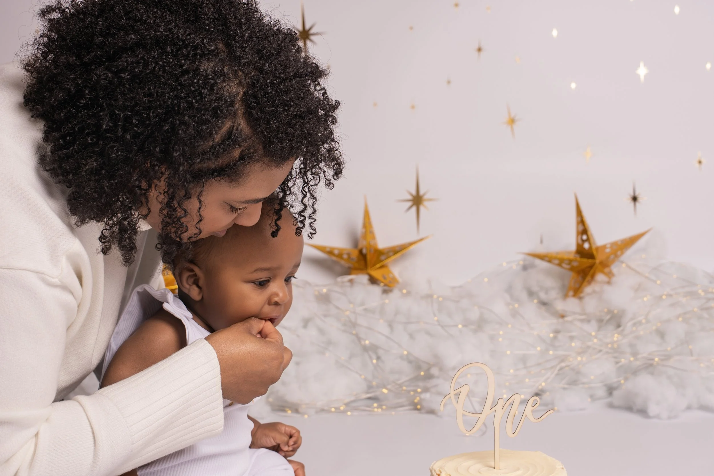 A woman and a small child celebrating a first birthday with a cake that has a wooden "One" topper. The background has white fluffy decorations, gold star-shaped ornaments, and string lights.