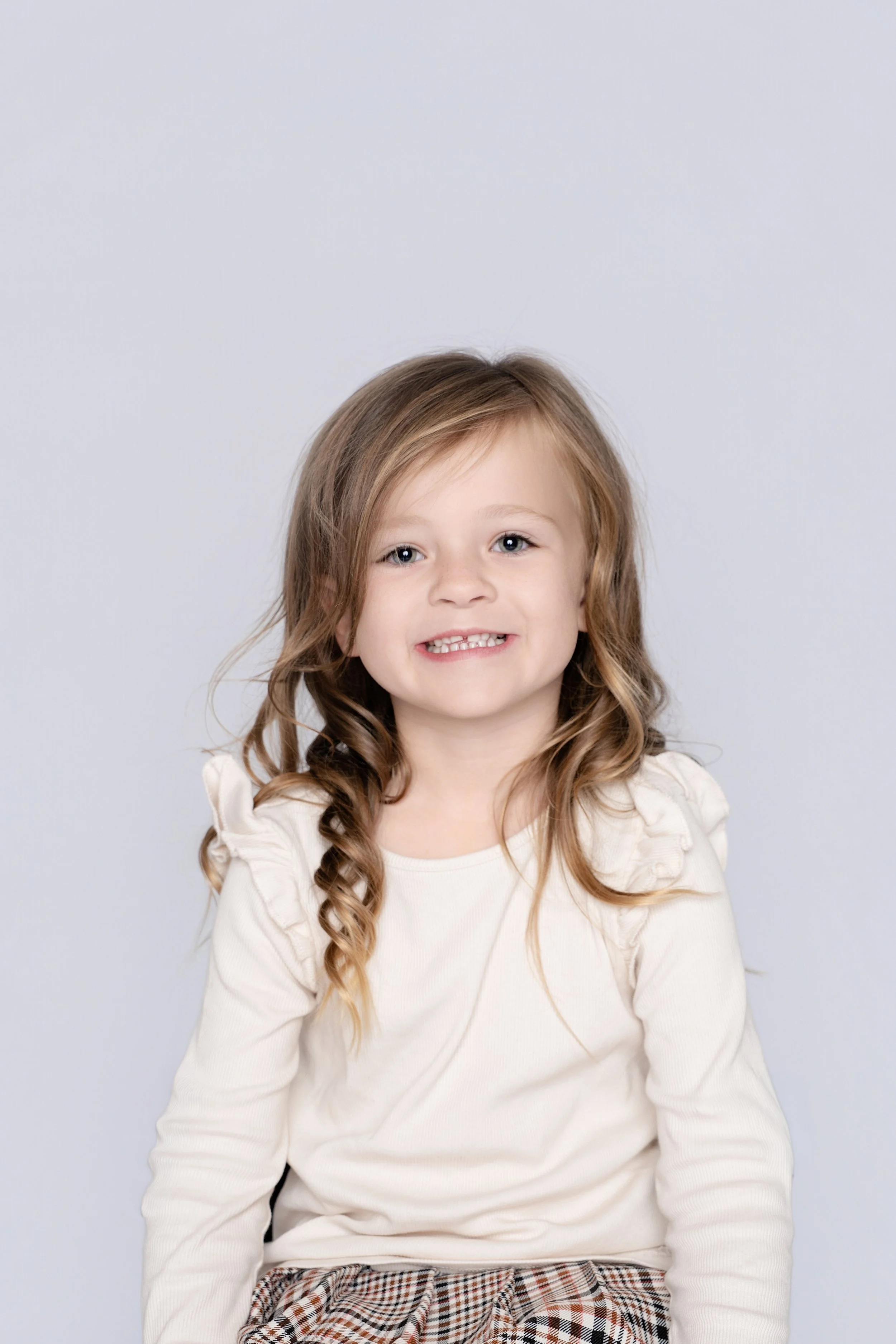 A young girl with long, wavy brown hair, smiling, wearing a cream-colored top with ruffled shoulders and plaid pants, sitting against a light gray background.