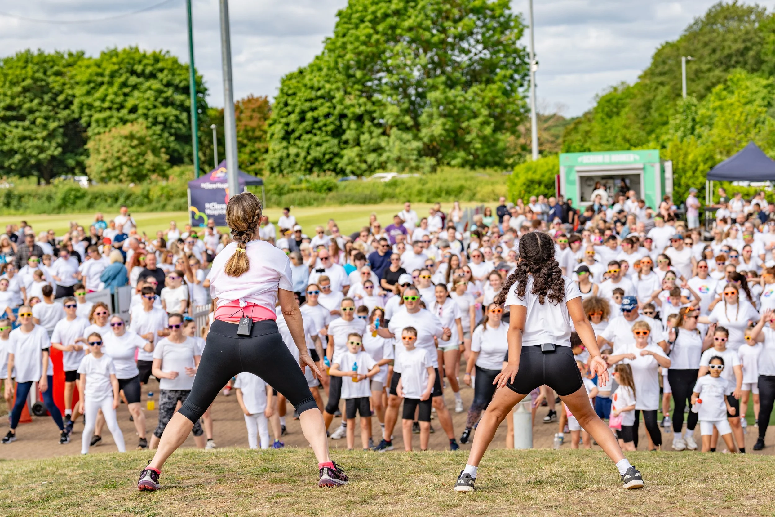 Two women leading an outdoor exercise class with a large crowd participating, all wearing white shirts, in a park with green trees and cloudy sky.