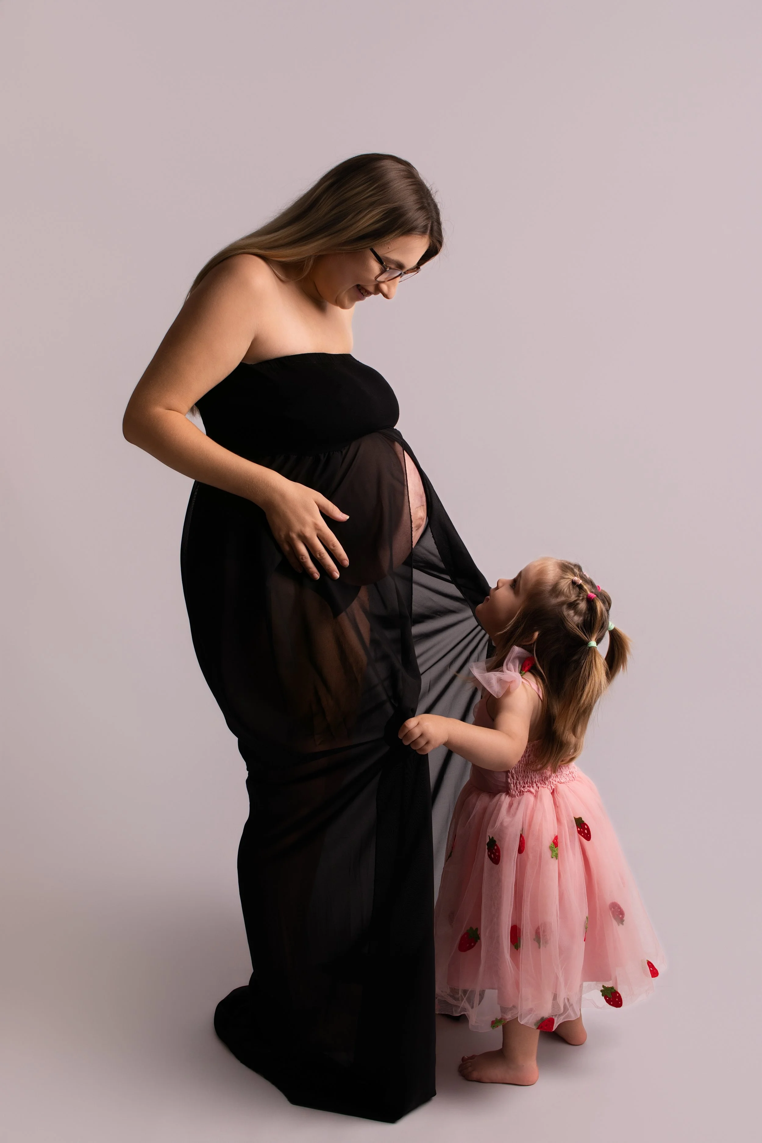 A pregnant woman in a black dress and glasses is smiling while her young daughter in a pink dress with strawberries pattern gently touches her belly and looks up at her.