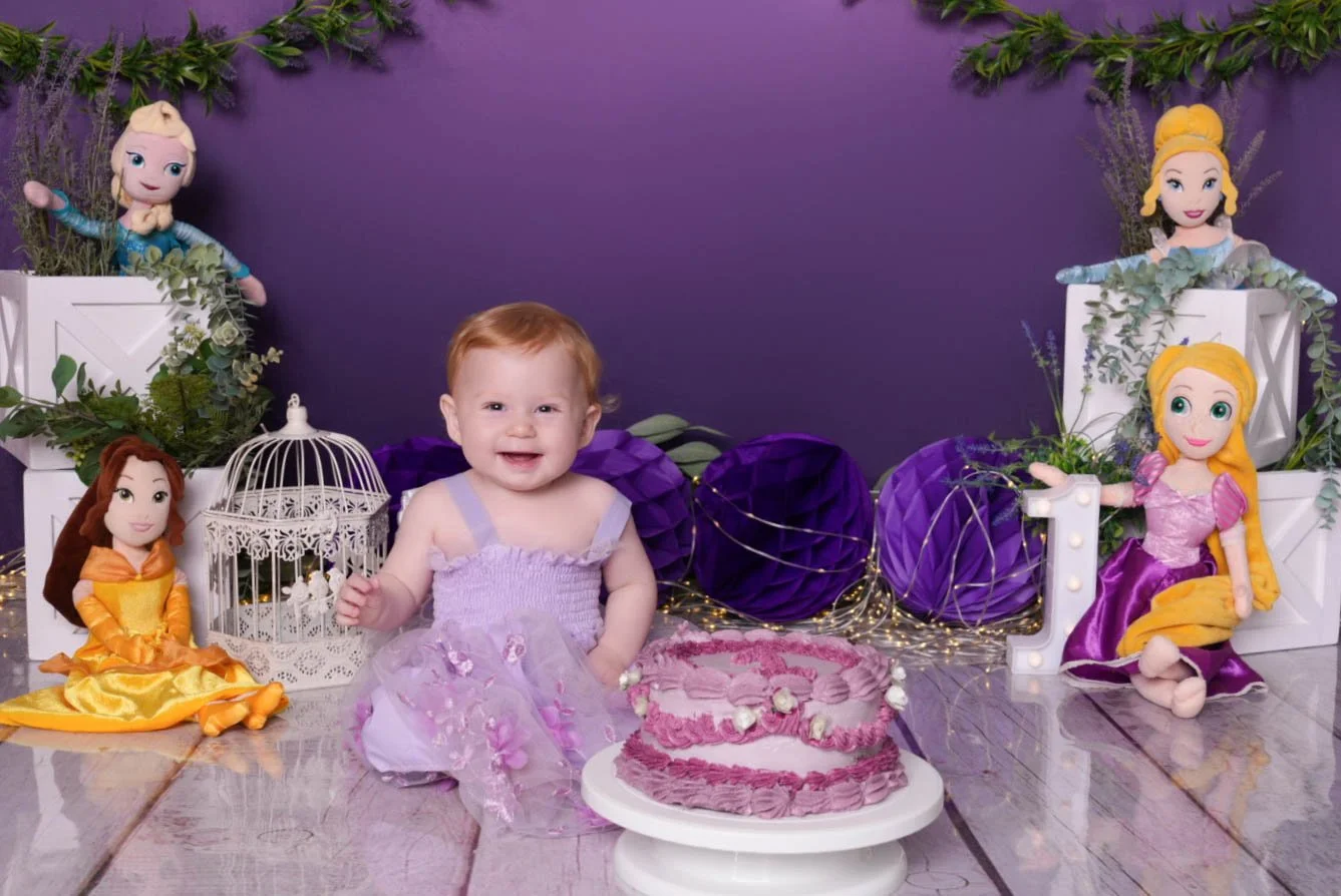Child's birthday celebration with a birthday cake, surrounded by plush Disney princess dolls, purple decorations, and a purple background.