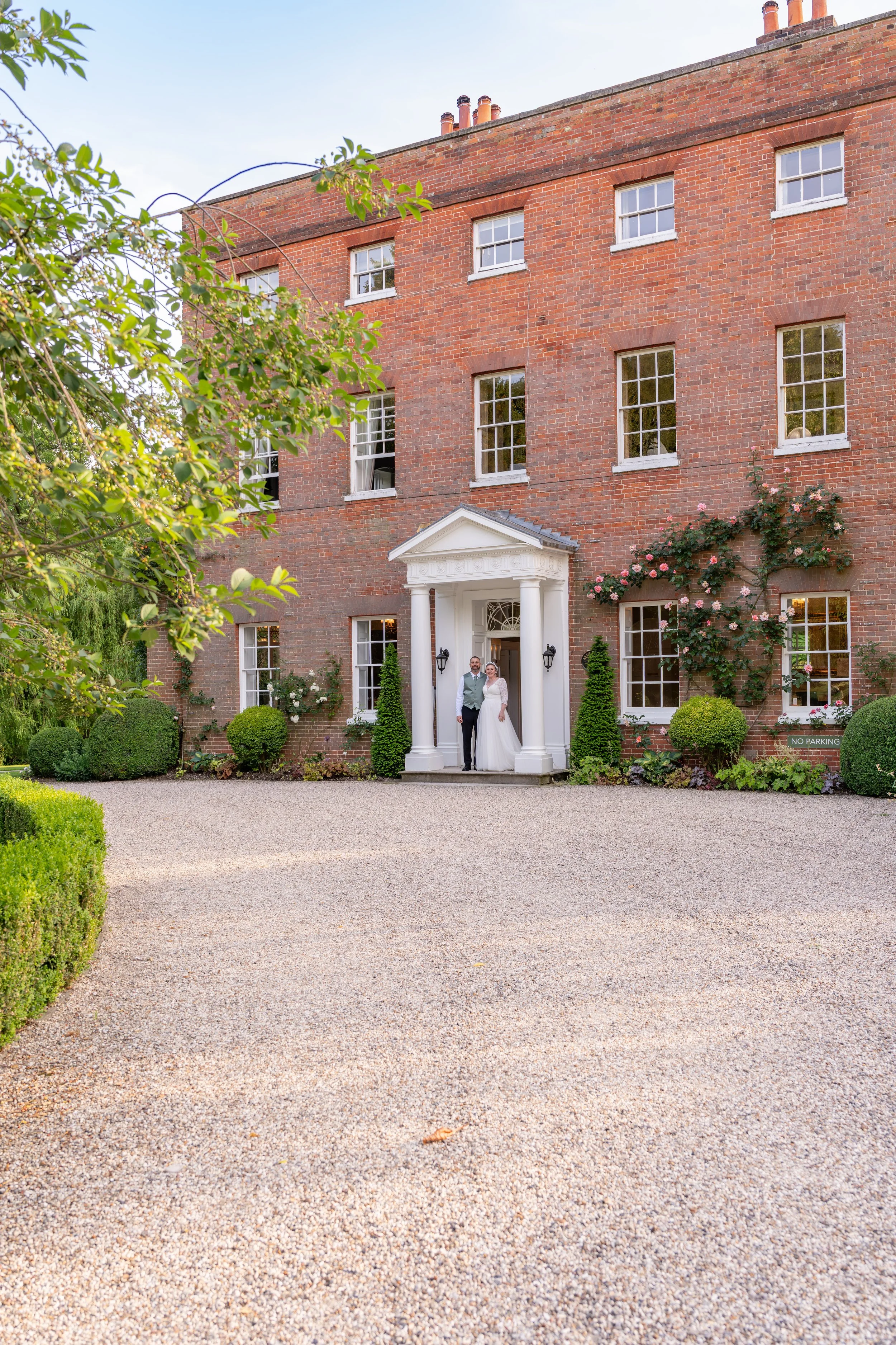 A bride and groom standing at the entrance of a large brick house on their wedding day.