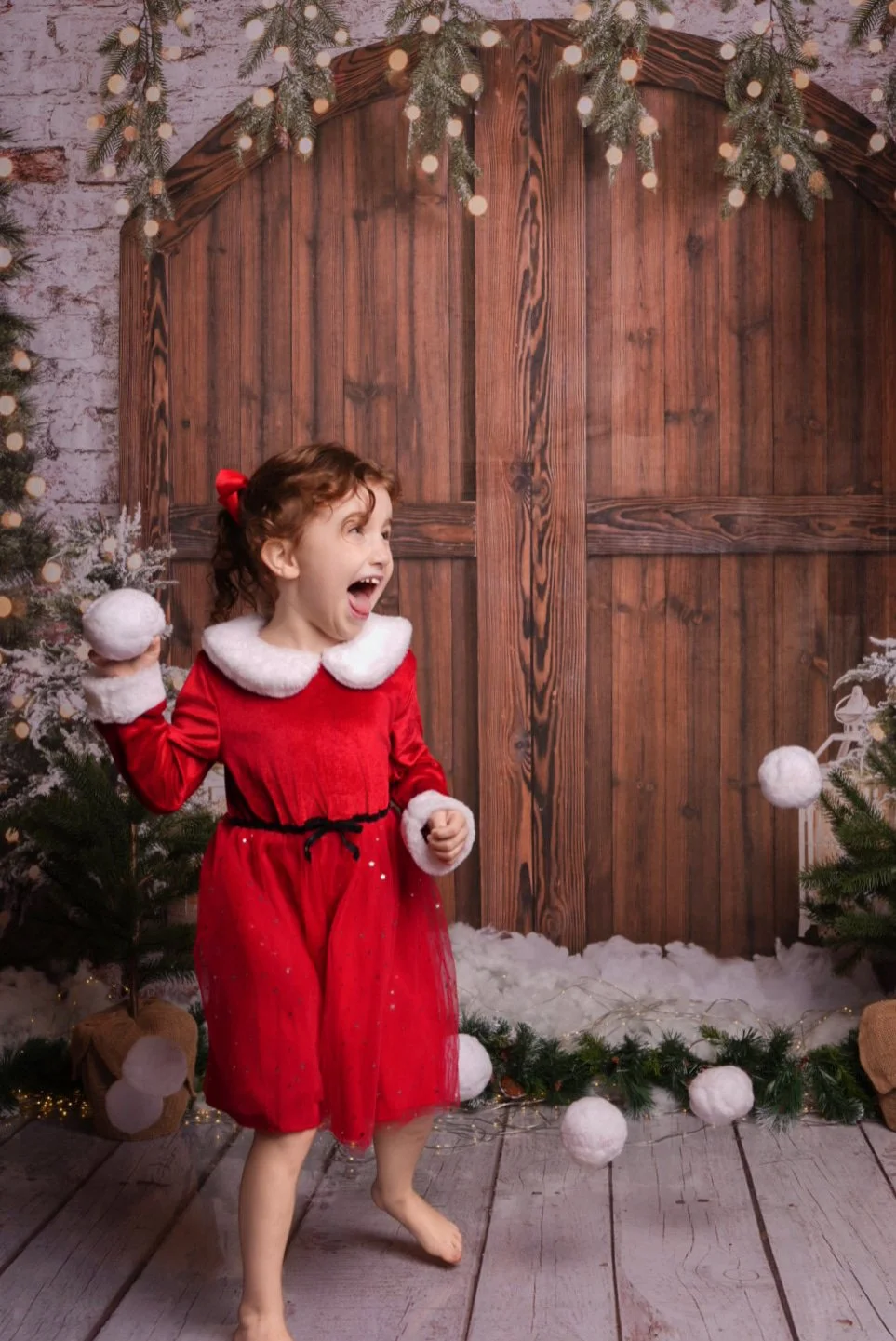 A young girl dressed in a red holiday dress with white fur collar and cuffs, smiling and holding a snowball, standing on a wooden floor in a Christmas setting with decorated trees, artificial snow, and string lights.