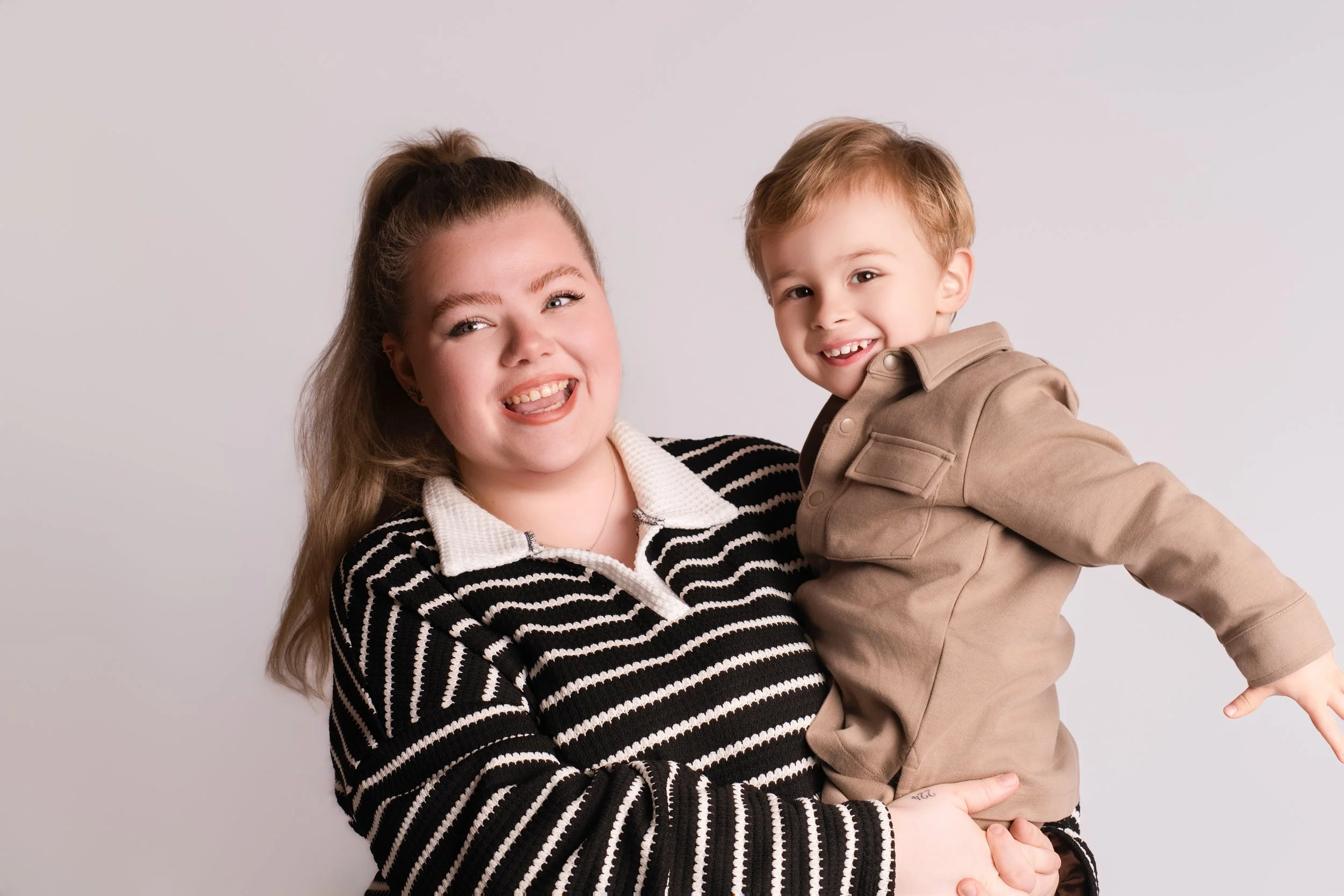 A woman holding a young boy, both smiling and looking at the camera, in front of a plain light gray background.