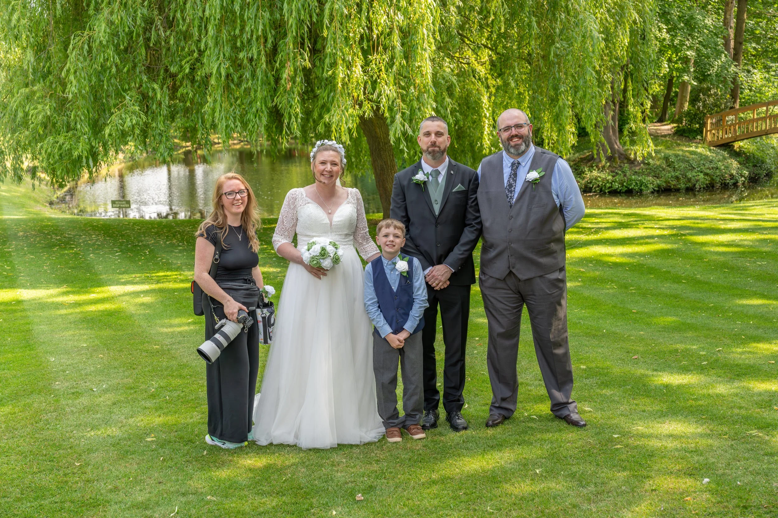 A wedding party standing on a grassy area near a pond, with trees and a small wooden bridge in the background. The bride is wearing a white gown holding a bouquet of white roses. The groom is dressed in a dark suit with a light green tie and boutonniere. A young boy, likely the ring bearer, is in a blue vest and tie. The woman on the left has a camera hanging from her neck, and the man on the right is wearing a gray vest over a blue shirt.