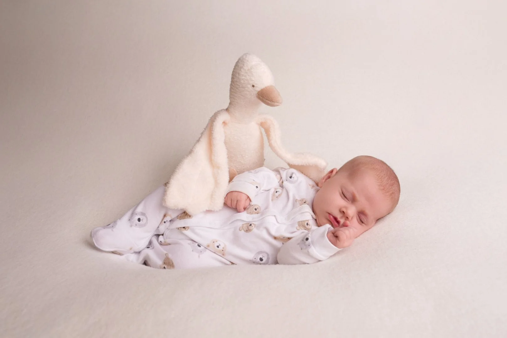 A sleeping baby in a white onesie with teddy bear prints, lying on a cream-colored surface, with a plush duck toy beside them.