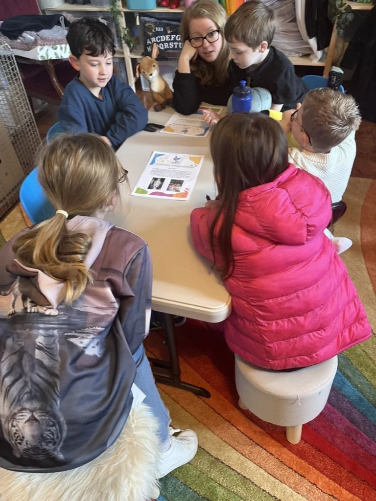 A group of children and a woman gathered around a table looking at a colorful sheet of paper with images and text. The children are seated and the woman is leaning in, engaged with the kids. The setting appears to be a classroom or a playroom with books, toys, and colorful shelves in the background.