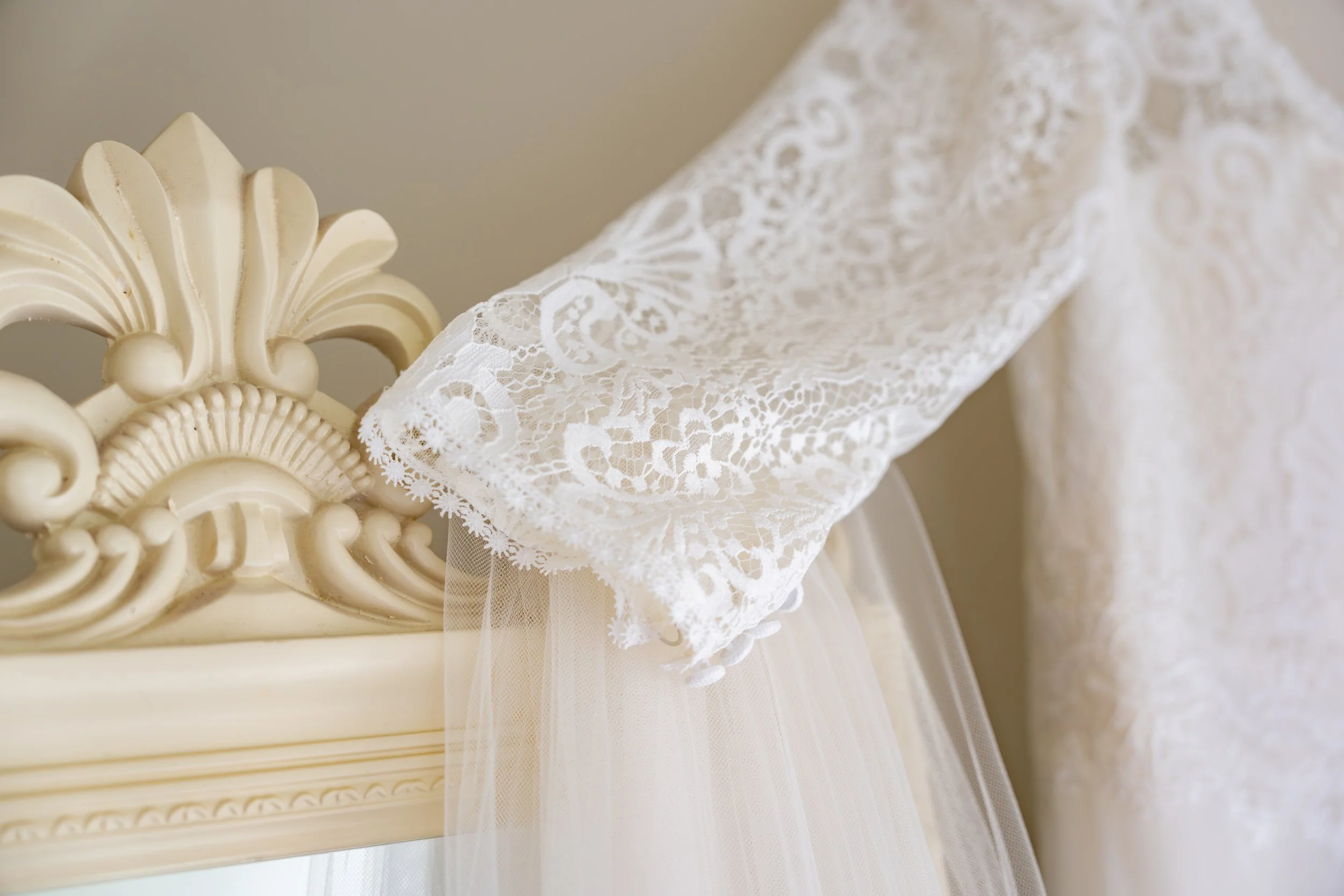 Close-up of a vintage white lace wedding dress with a tulle train, hanging on an ornate cream-colored vintage headboard.