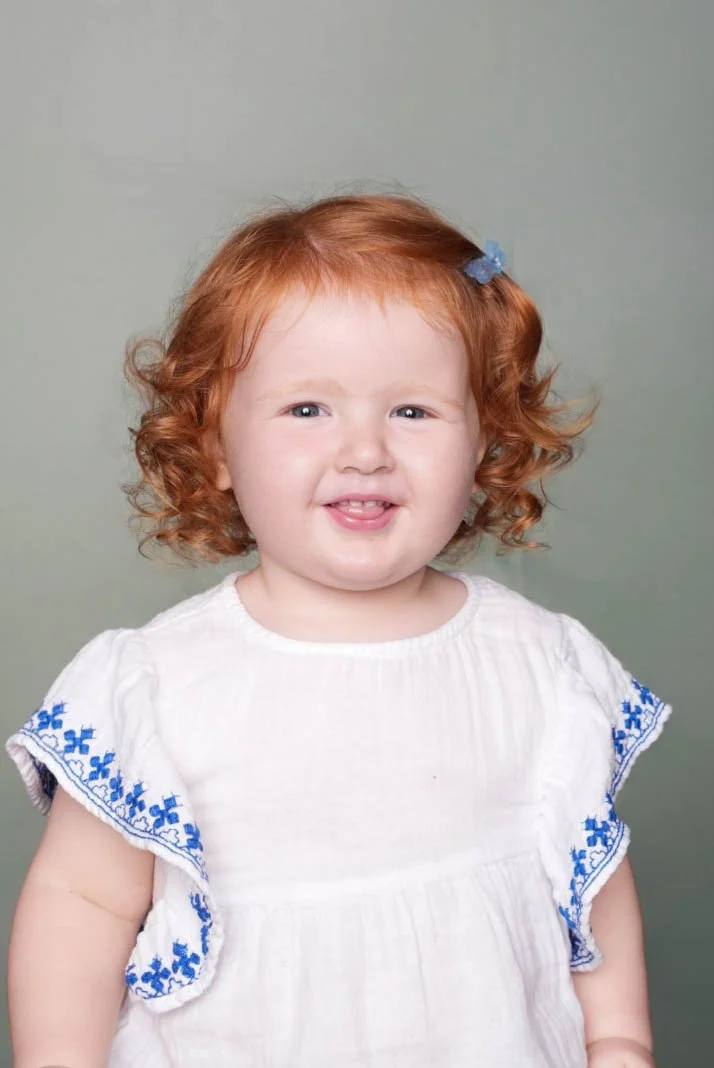 A young girl with curly red hair and a small blue bow, wearing a white dress with blue embroidery on the sleeves, smiling and sticking out her tongue, against a plain background.