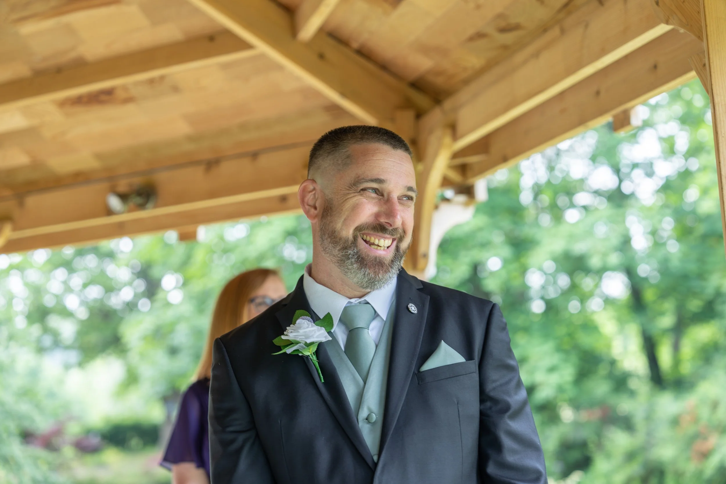 A smiling man dressed in a black suit with a light gray tie and pocket square, wearing a white boutonniere, at an outdoor wedding. There is a woman with red hair and glasses standing behind him, under a wooden shelter with green trees in the background.