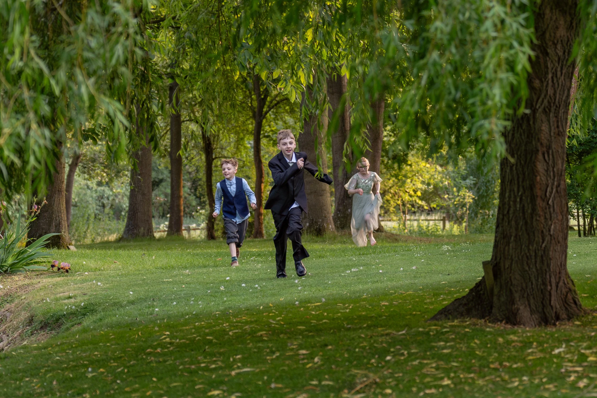 Three children in formal attire running through a lush green park surrounded by tall trees.