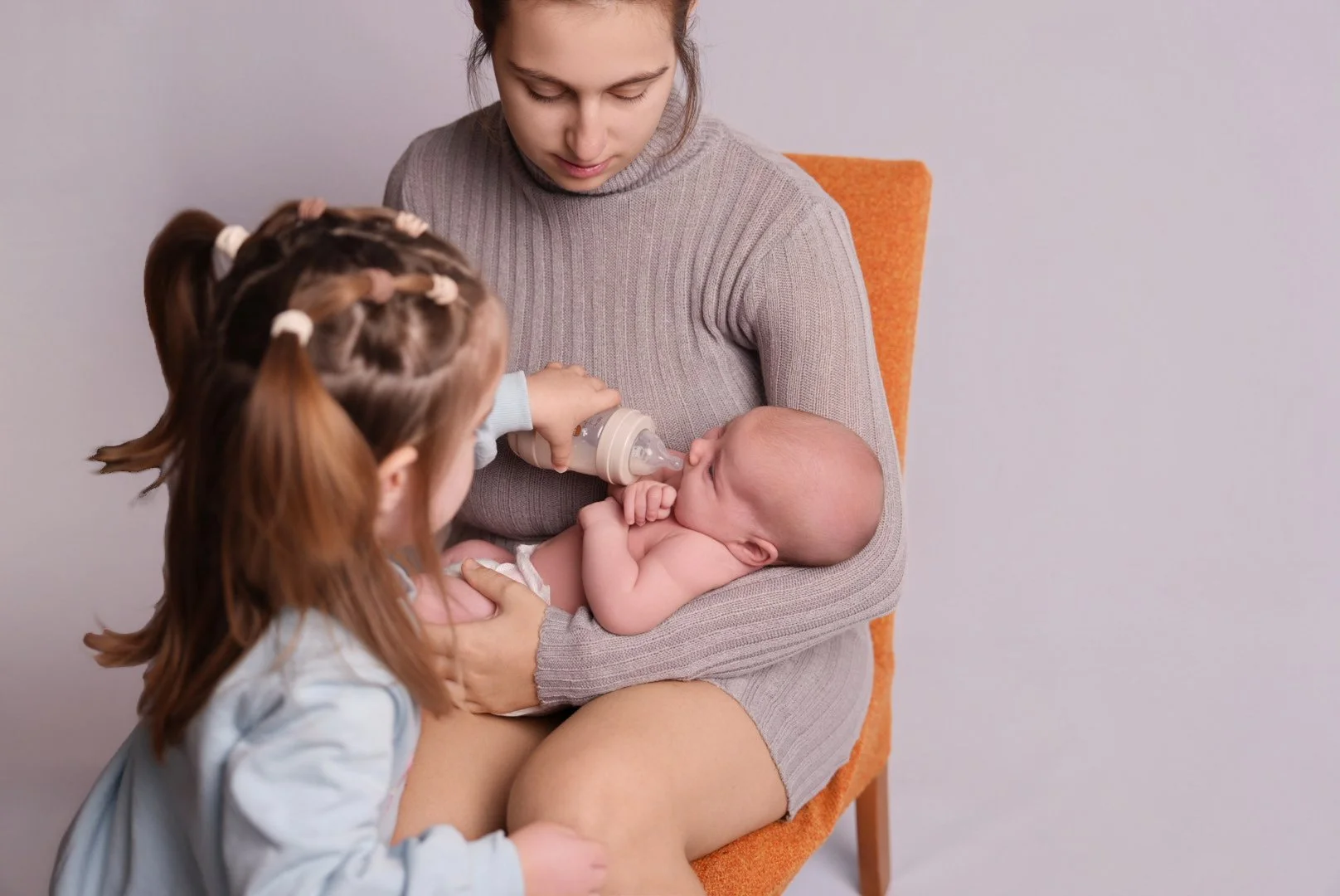 A woman holding a baby on her lap, a young girl feeding the baby with a bottle, all sitting in an orange chair against a plain light background.