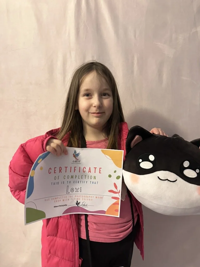 Young girl with brown hair in a pink jacket holding a colorful certificate of completion and a large plush black and white cat toy, standing against a plain wall.