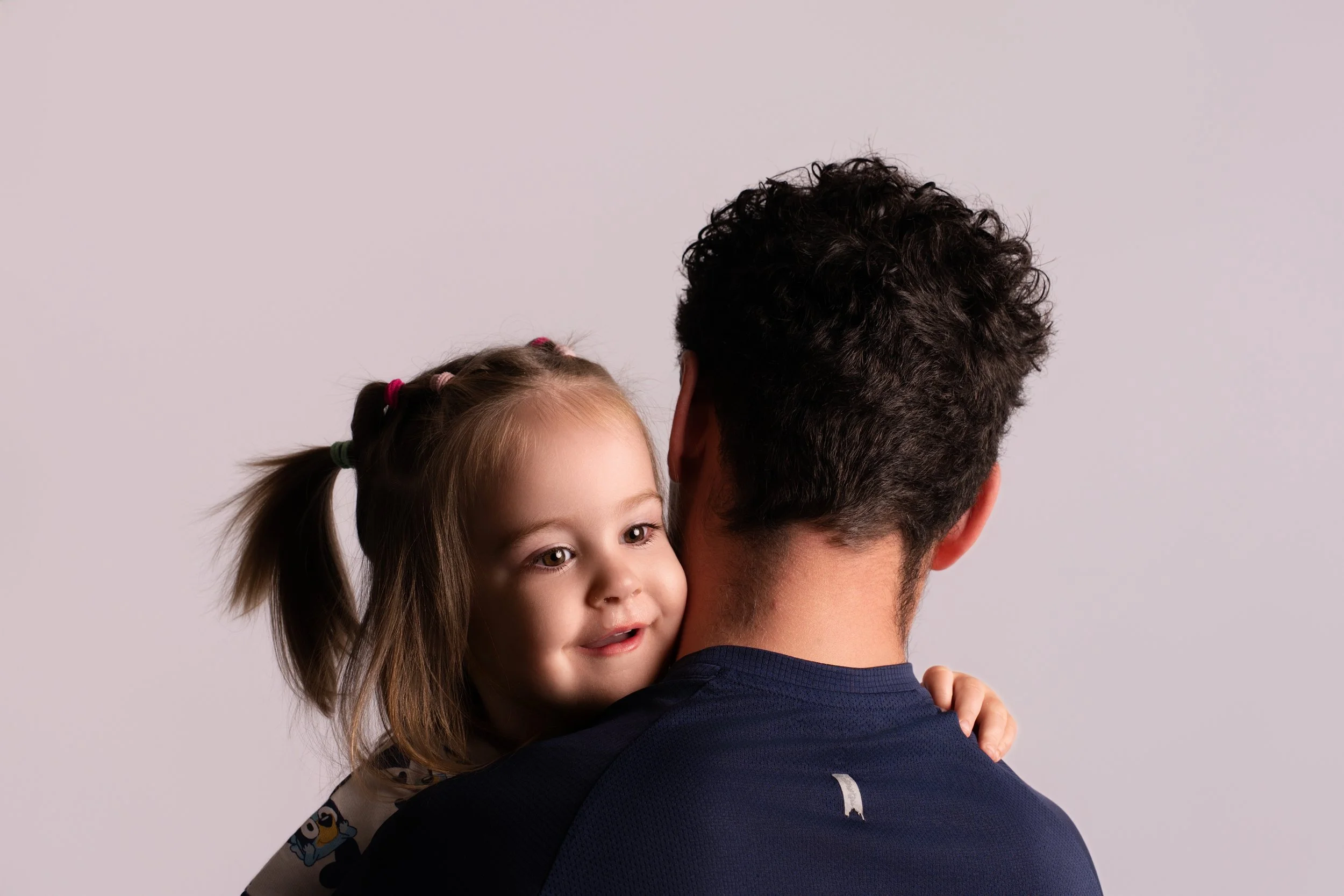 A man holding a little girl as she peeks over his shoulder smiling