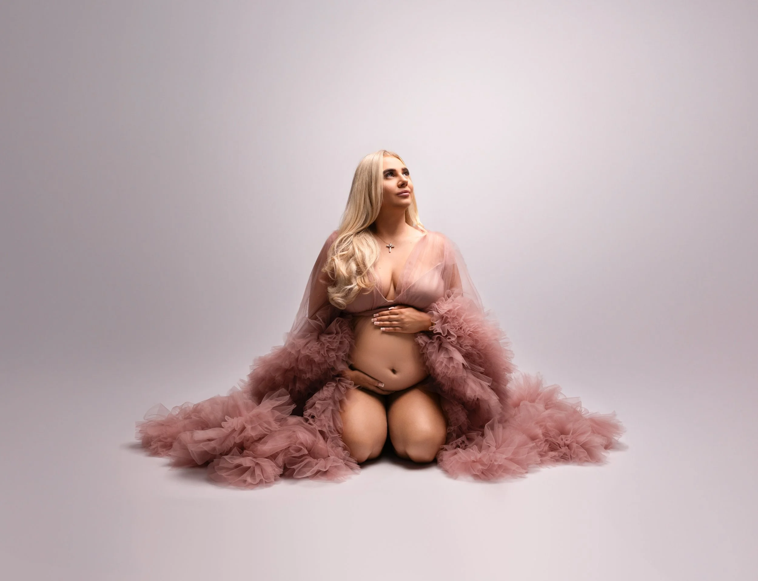 A pregnant woman kneeling on the floor, wearing a sheer pink gown with ruffled tulle around her legs, looking upwards with one hand on her belly and the other resting on her chest, against a plain white background.
