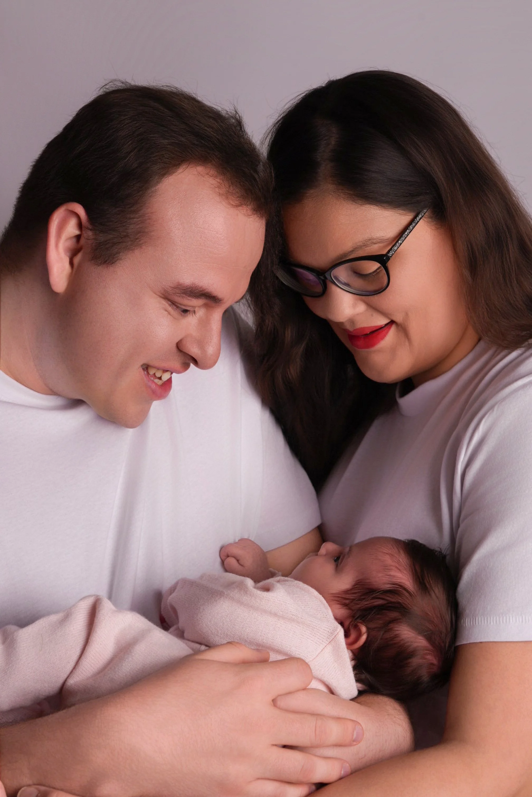 A couple holding a newborn baby, smiling, close together with their heads touching, against a plain background.