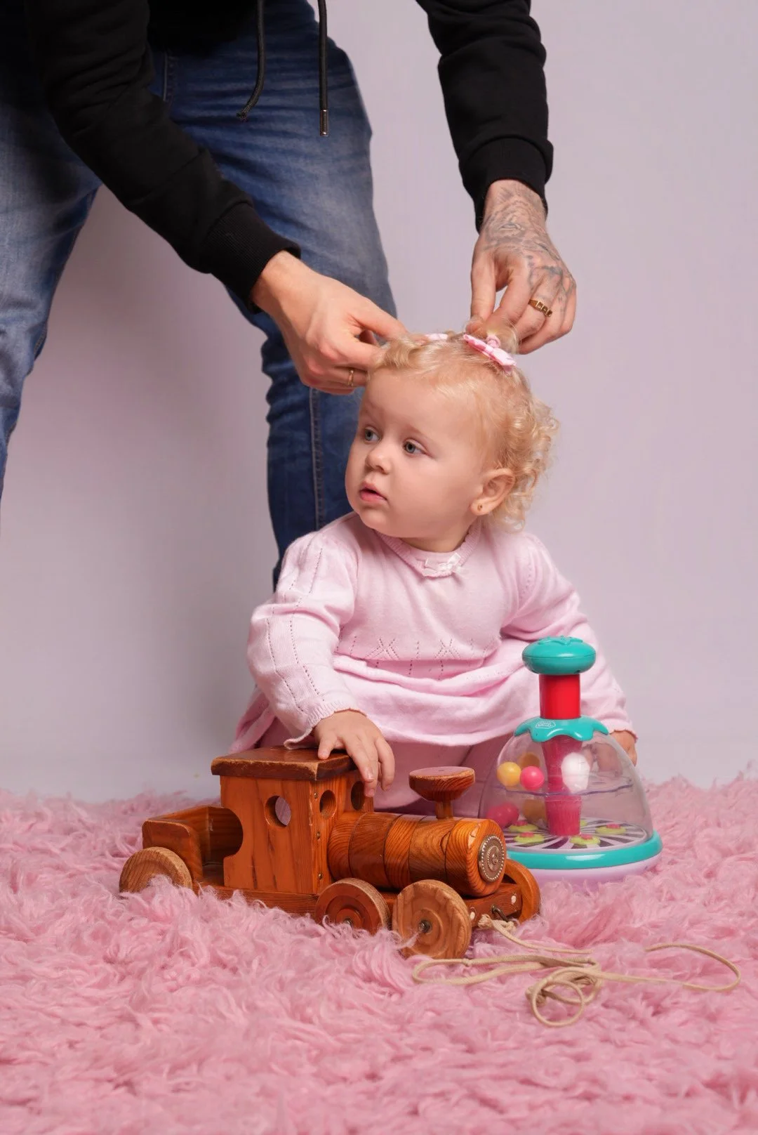 A young girl with curly blonde hair in a pink dress sitting on a pink fluffy rug, playing with a wooden toy train and a colorful spinning top. An adult is fixing her hair, visible from the waist down, wearing blue jeans and a black hoodie.