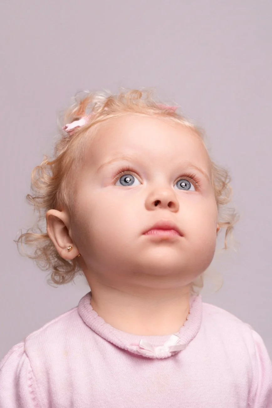 A young girl with curly blonde hair, blue eyes, and small earrings, looking upward with a neutral expression. She is wearing a light pink shirt.