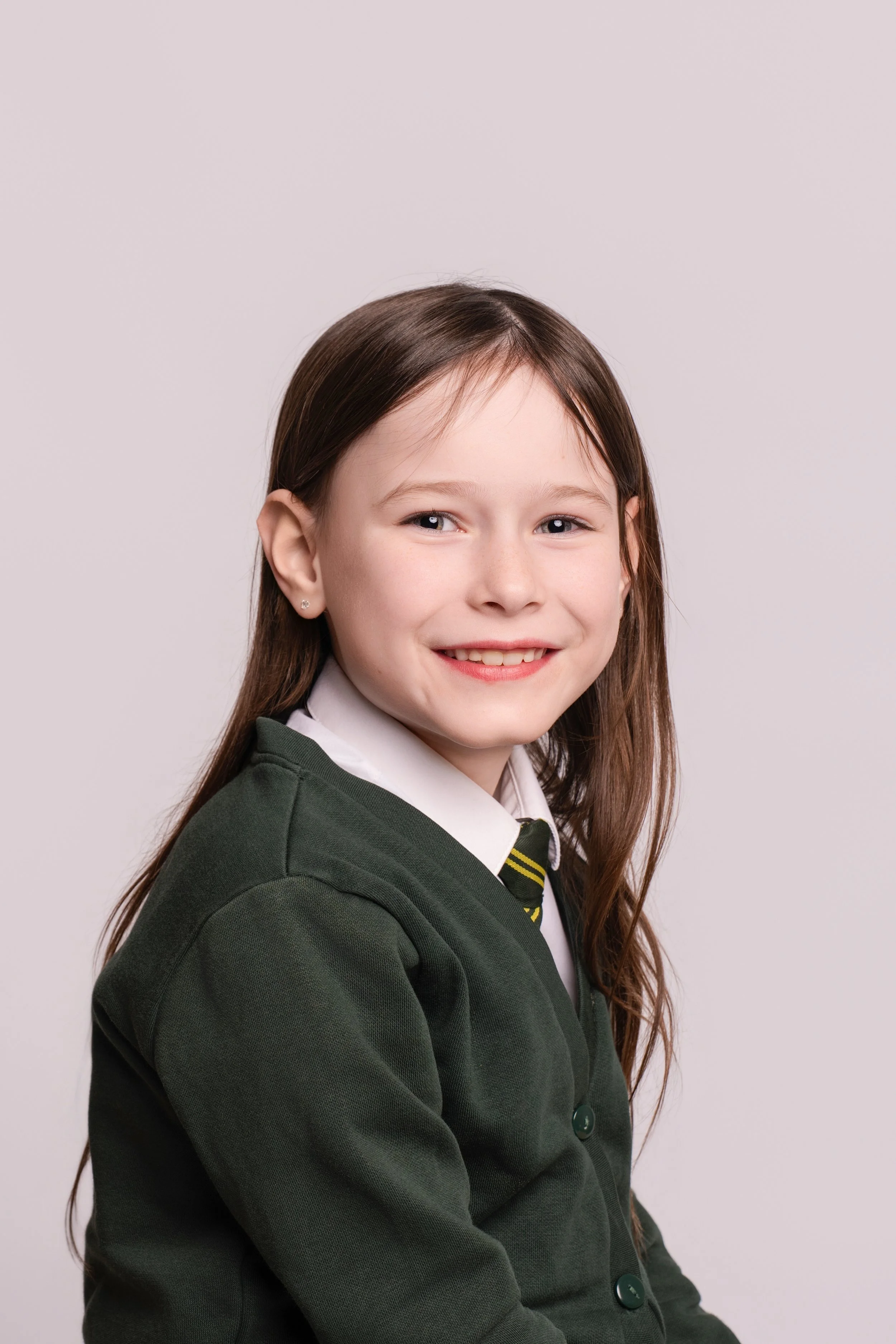 A young girl with long brown hair, smiling, dressed in a white shirt, black and yellow striped tie, and dark green school sweater, posing against a plain light background.