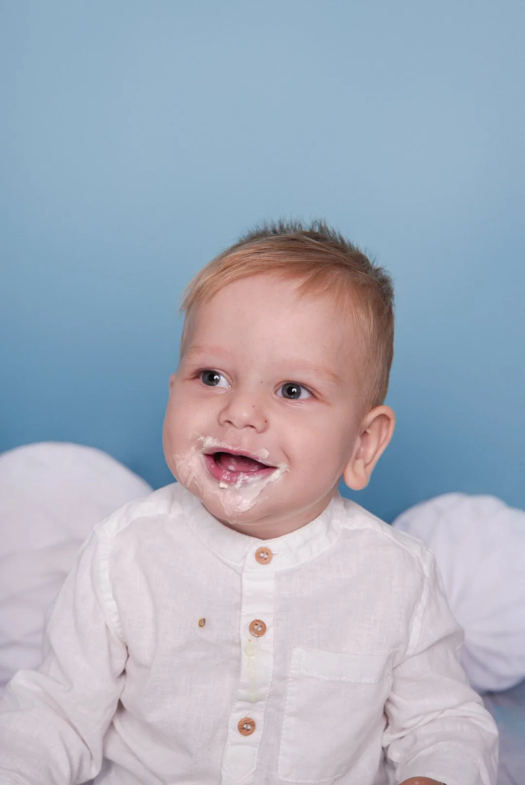 Smiling young boy with a messy face, wearing a white shirt, sitting against a blue background.