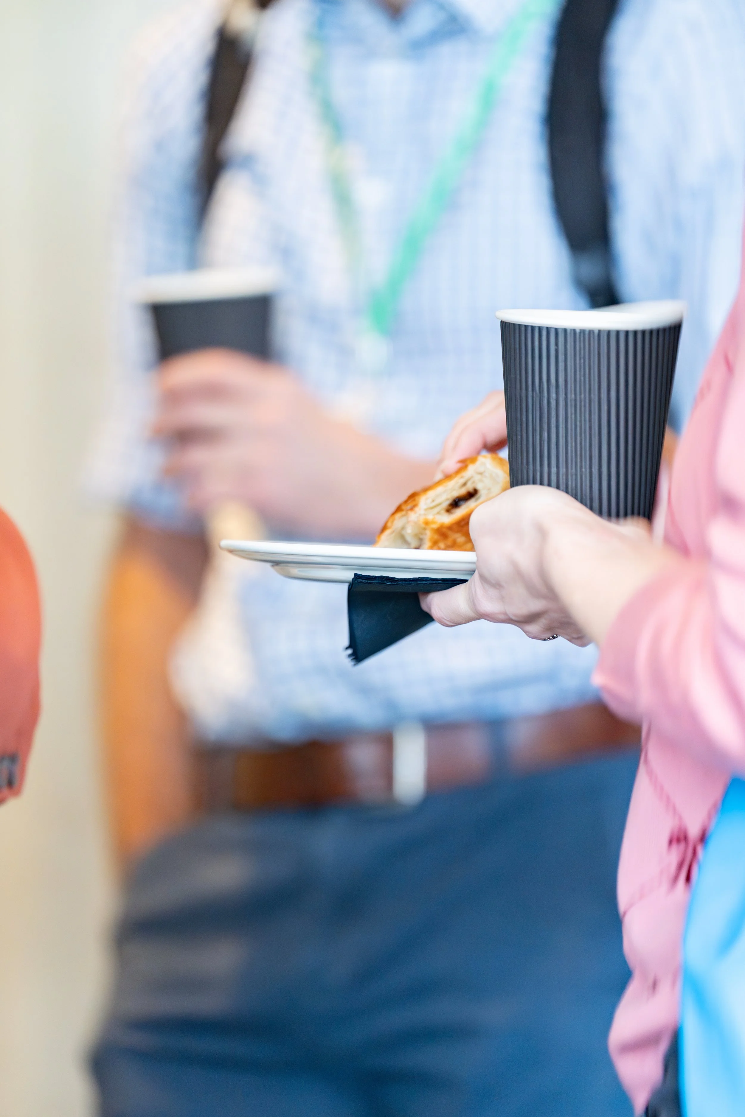 Close-up of a person holding a paper plate with a pastry and a paper cup, during a social event or conference.