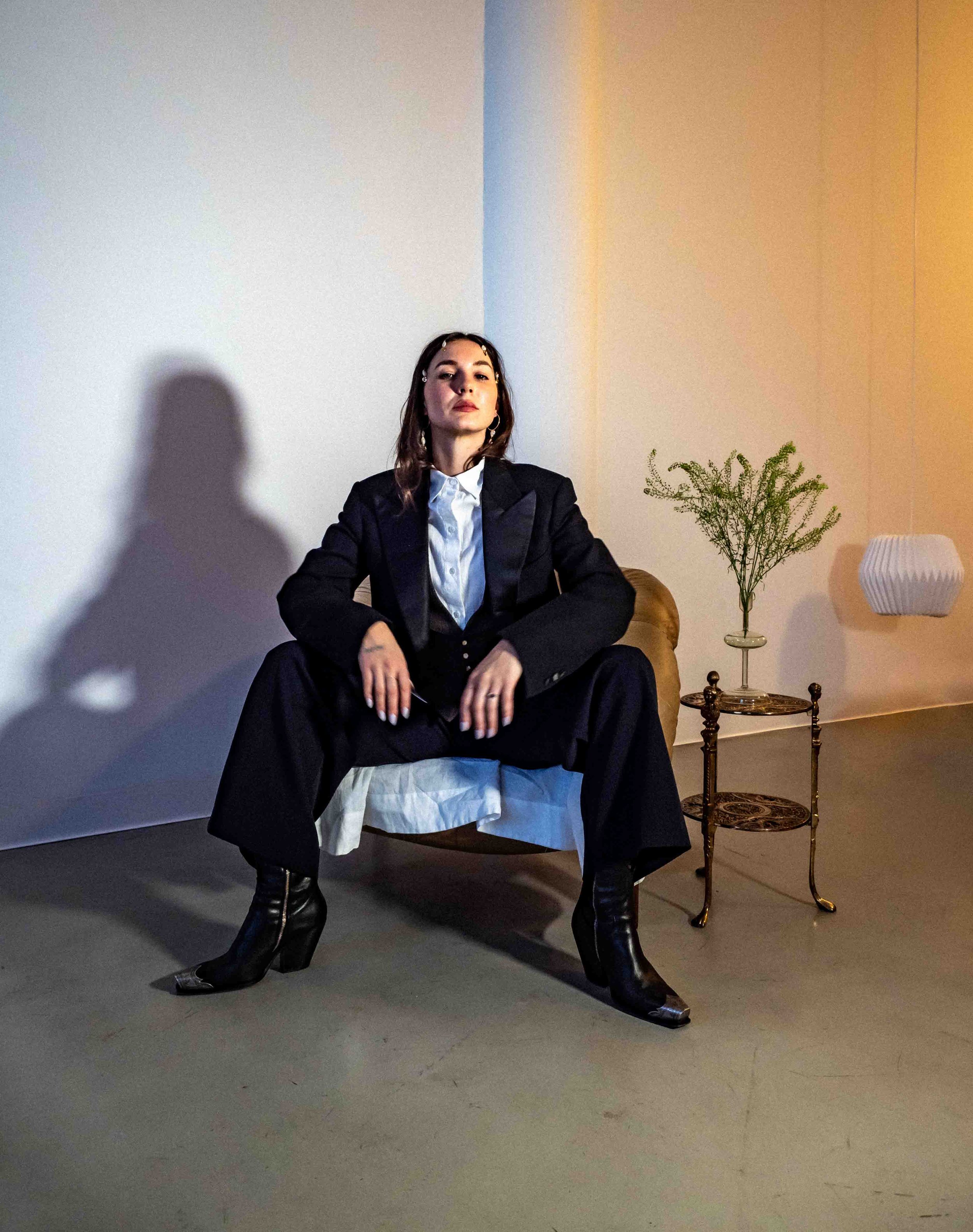 A woman in a black suit sitting on a chair, with her legs spread and hands resting on her knees, in a room with plain walls, a small side table with a plant, and a lamp.