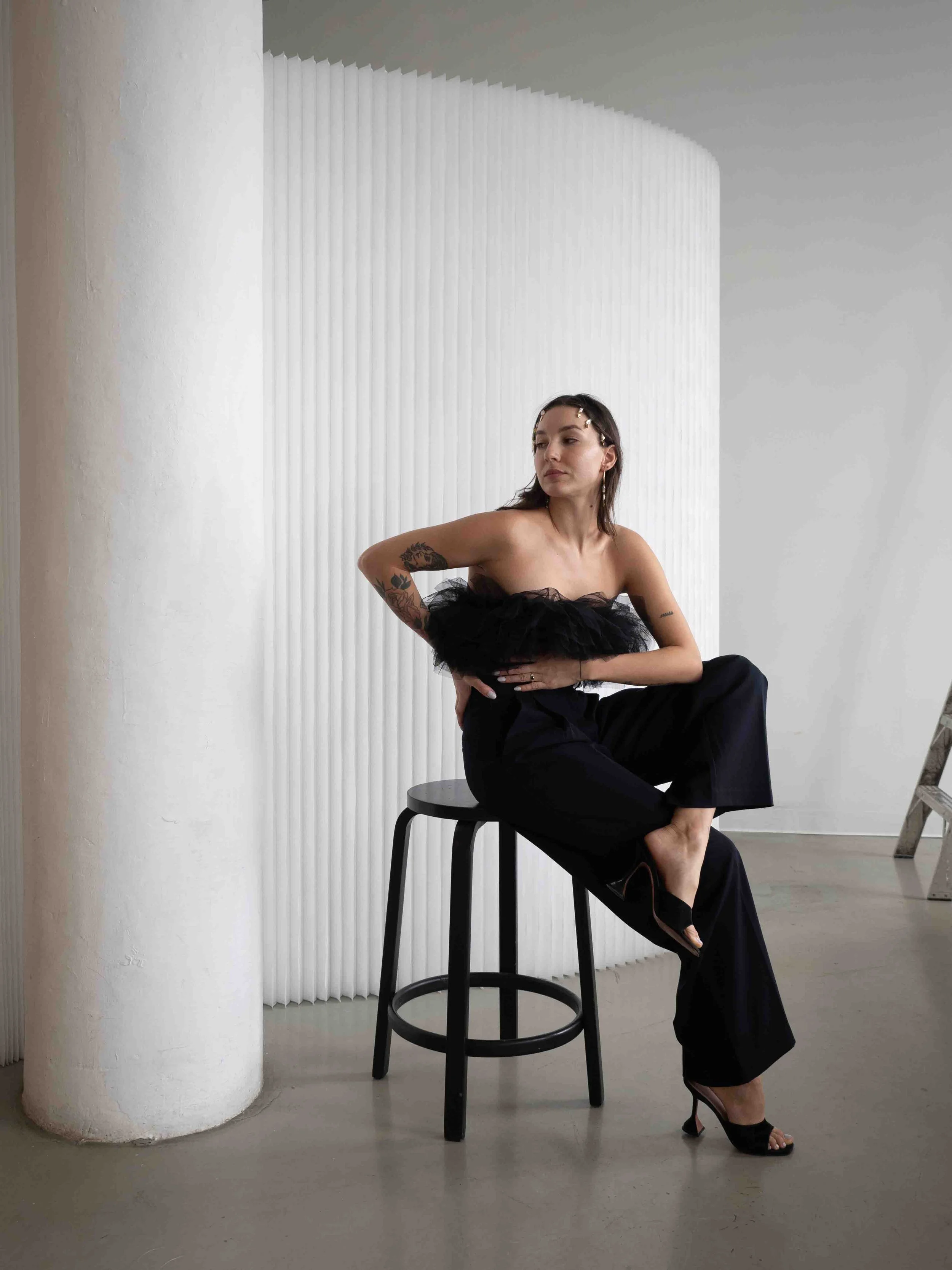 A woman with tattoos in a black strapless top and wide-leg black pants, wearing black high heels, sits on a black bar stool in front of a textured white wall. She is looking to the side with a neutral expression.