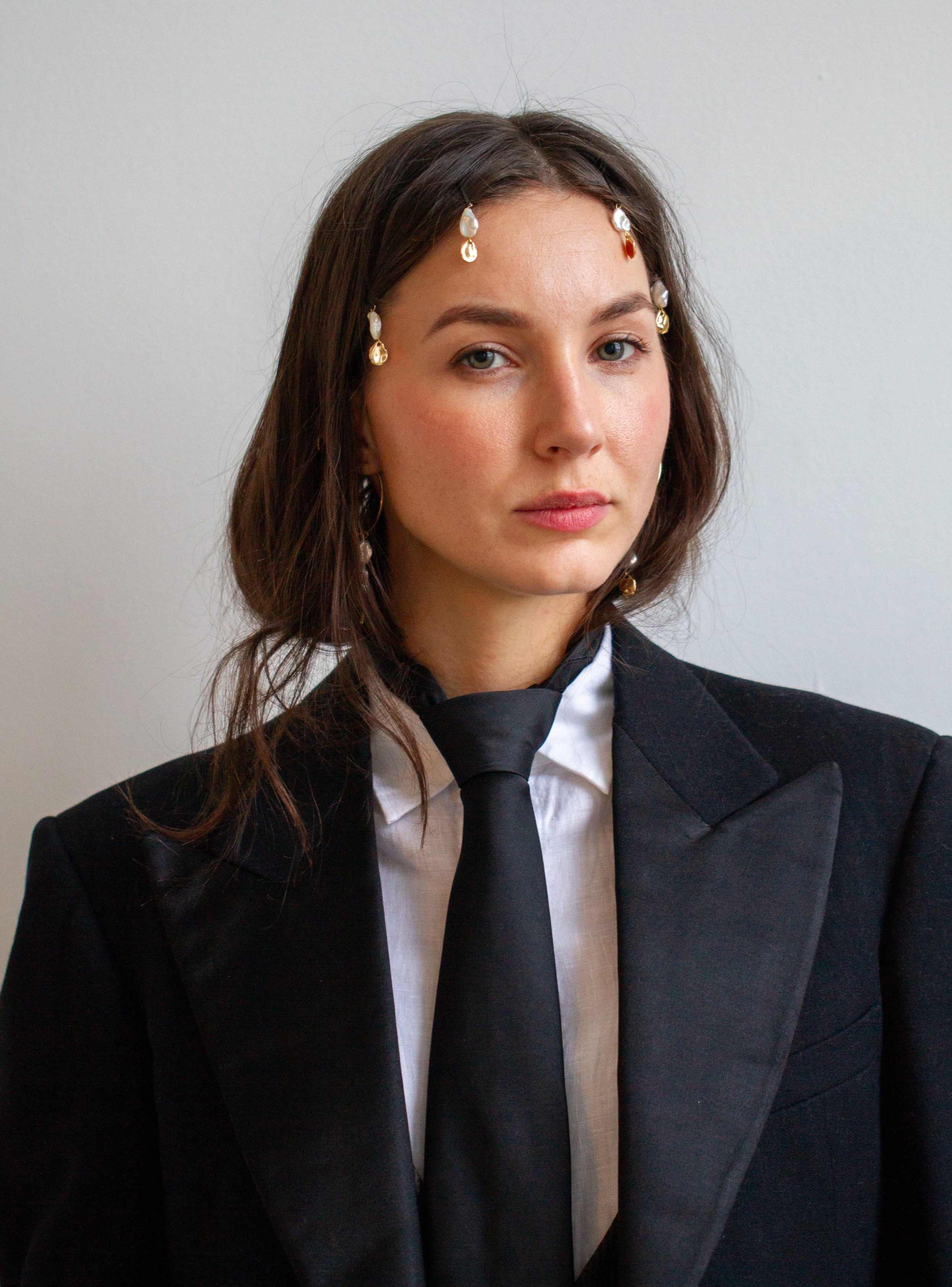 A woman with shoulder-length brown hair and light skin wearing a black suit jacket, white shirt, black tie, and pearl and gemstone earrings. She is standing against a plain light background.