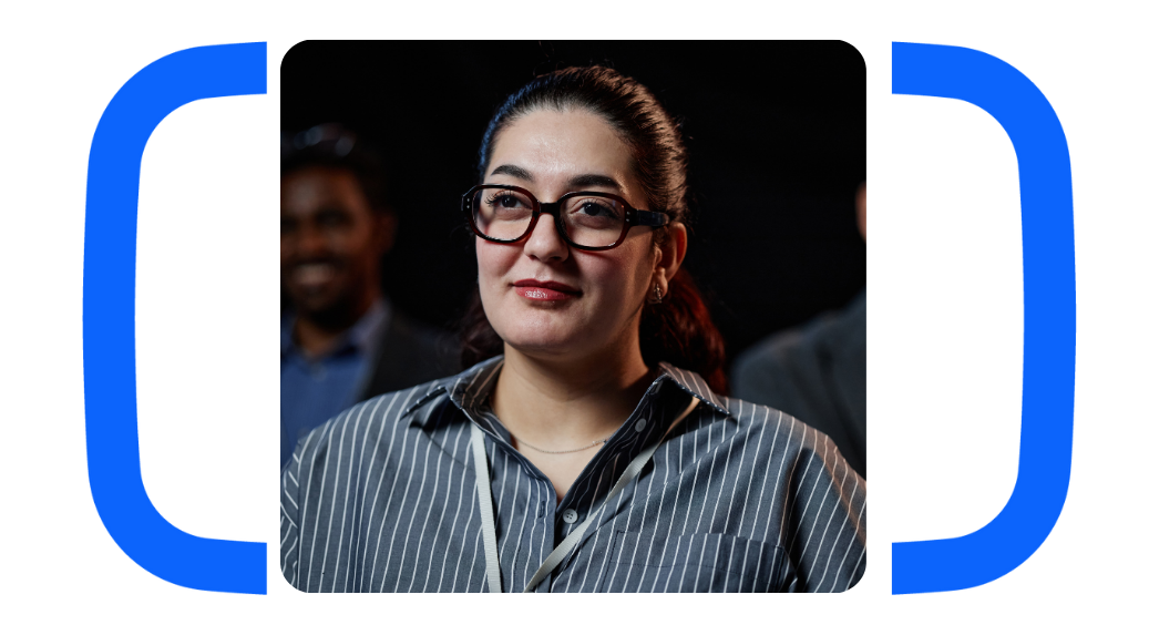 A woman with glasses and a striped shirt looking thoughtfully, with blurred people in the background.