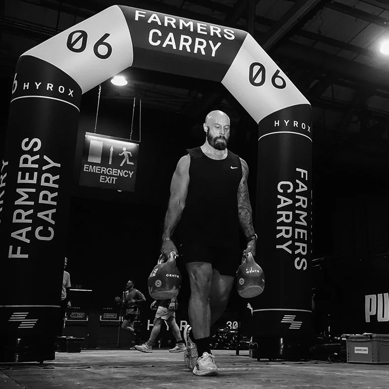 A man walking through a fitness event archway labeled 'Farmers Carry' and 'Hyrox' while holding sandbags, with other participants and an emergency exit sign in the background.