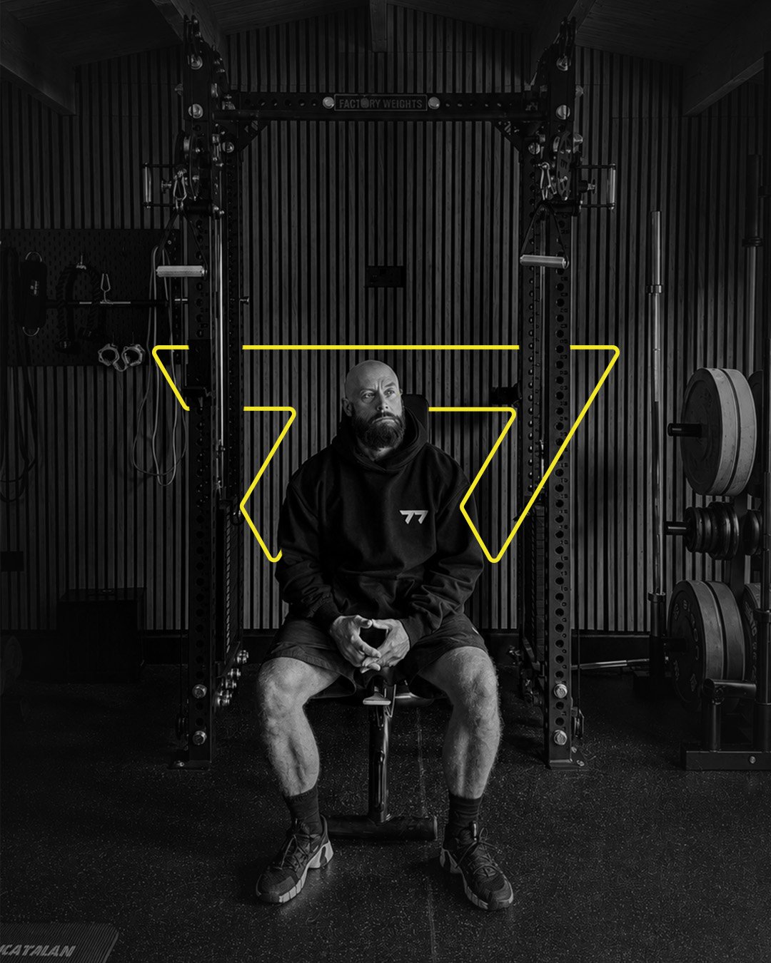 A man with a beard and shaved head sitting in a gym, surrounded by workout equipment including weight plates and a squat rack, with yellow lines drawn over his body.