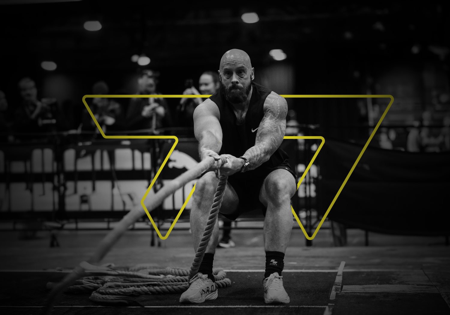 Black and white photo of a muscular man with tattoos on his right arm, wearing a black sleeveless shirt and shorts, participating in a tug-of-war, pulling on a thick rope on a gym floor with an audience watching in the background.