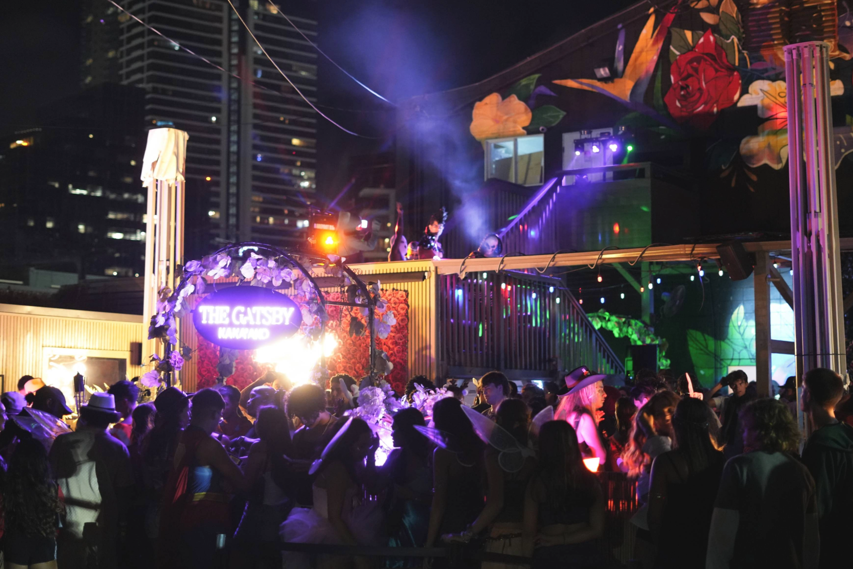 Nighttime scene at a club or event with a crowd of people, colorful lights, DJ booth, and floral decorations, with a sign reading 'The Gatsby Kamado'