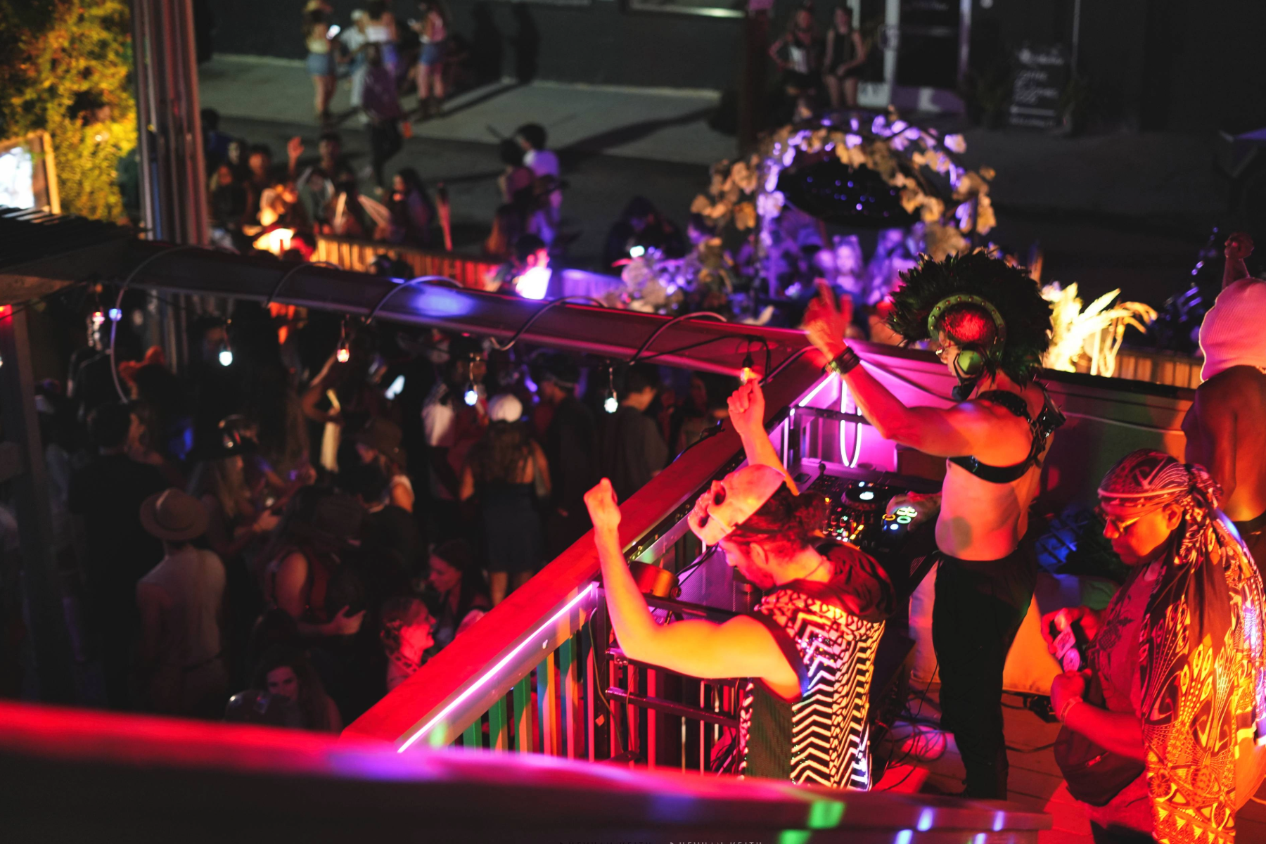Nighttime scene at a lively outdoor party with a DJ wearing a feathered headdress and sunglasses, dancing behind a DJ booth with colorful neon lights, while a crowd dances and socializes around them.