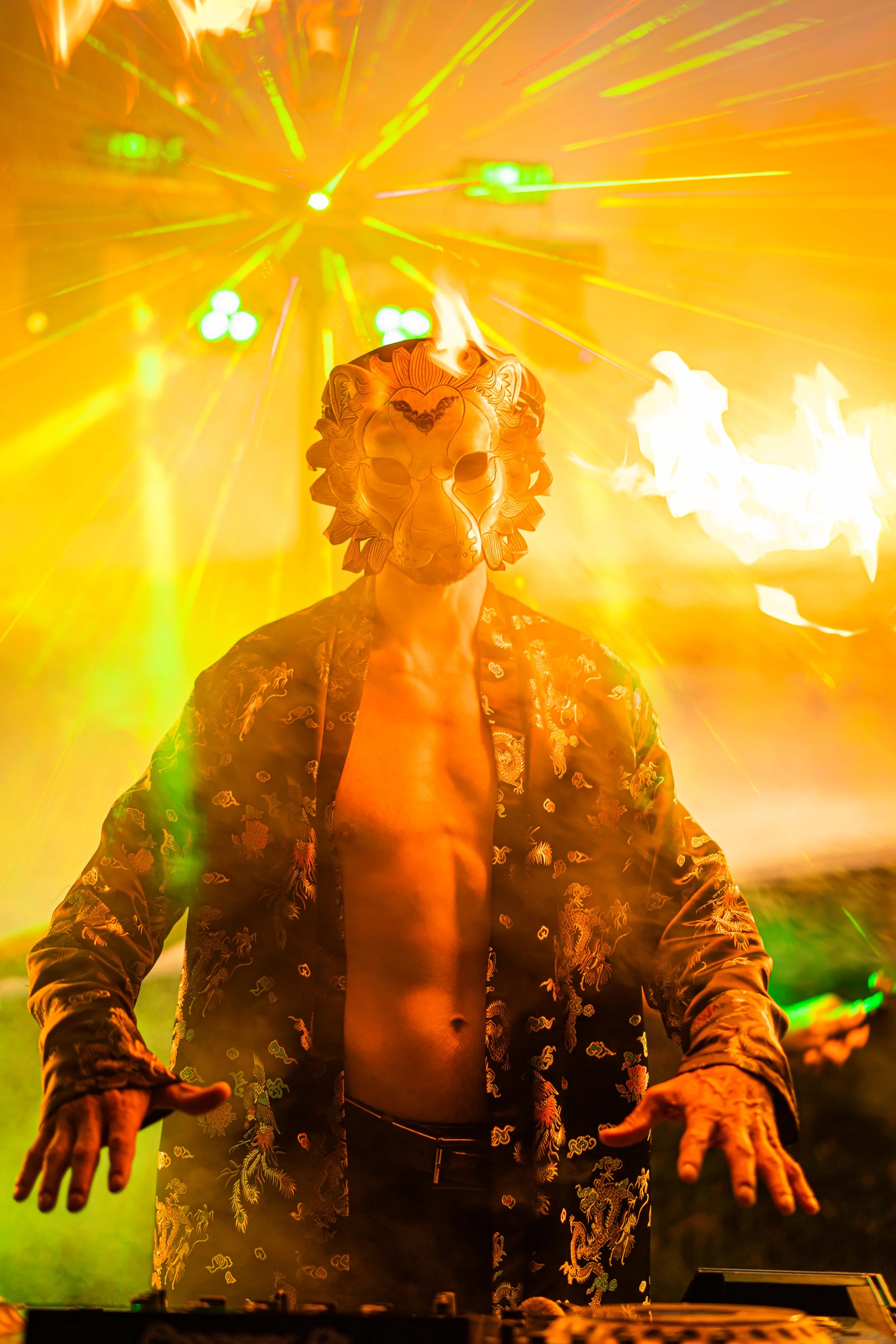 A person wearing a lion mask and a patterned shirt performing as a DJ at a concert or party, with bright yellow and green stage lights and open flames in the background.