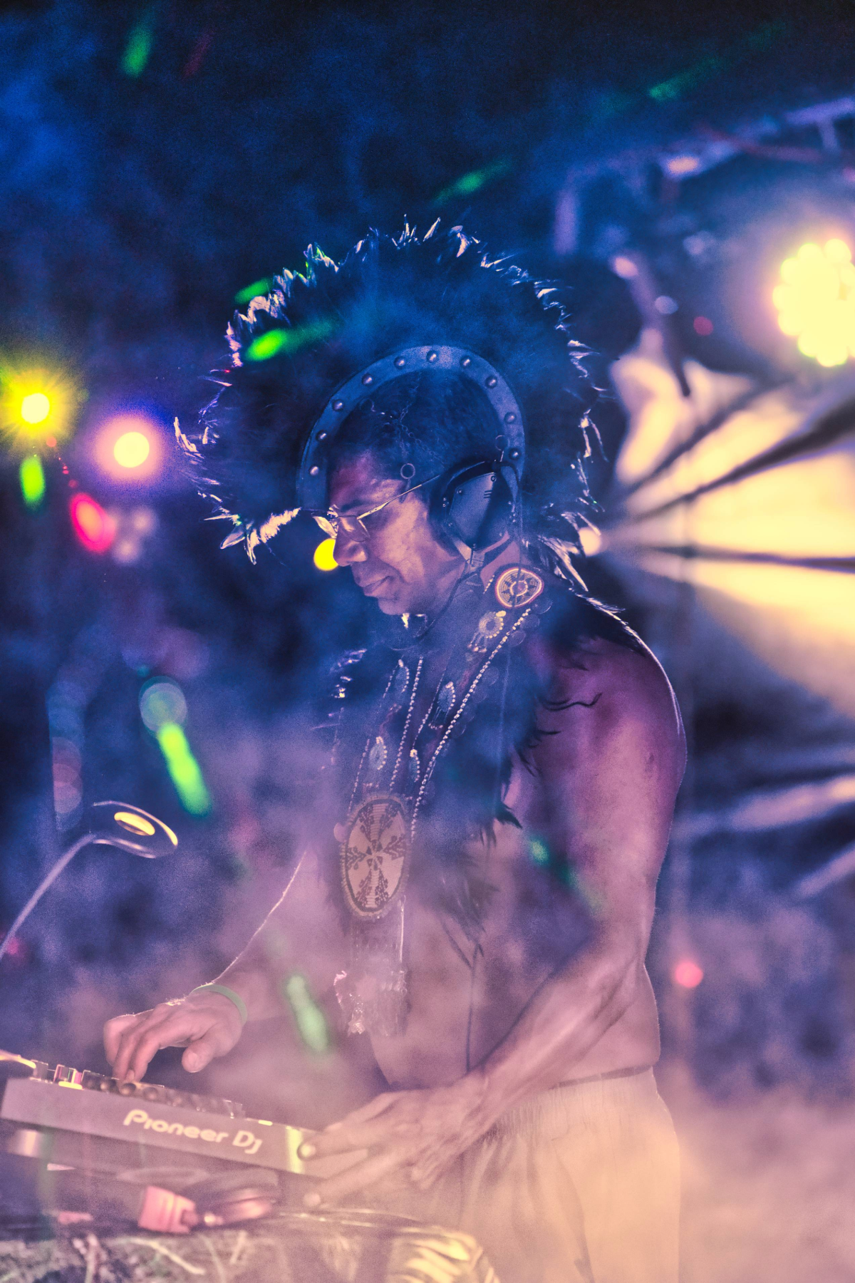 A DJ with an elaborate feathered headdress and necklaces performing with a Pioneer DJ mixer at a vibrant, colorful event with stage lights.